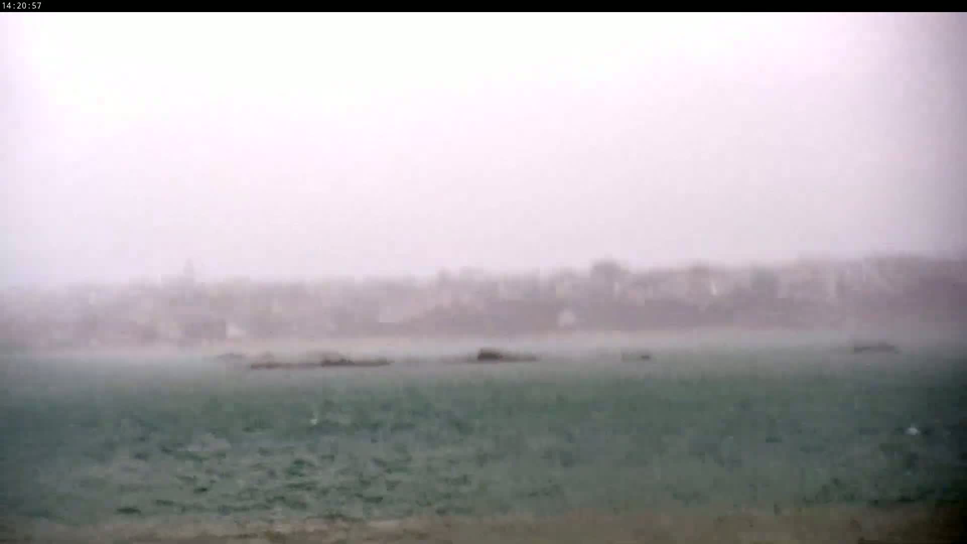 A hazy, rain-swept coastal scene shows a town in the distance and a choppy sea with small rocks partially submerged in the foreground.