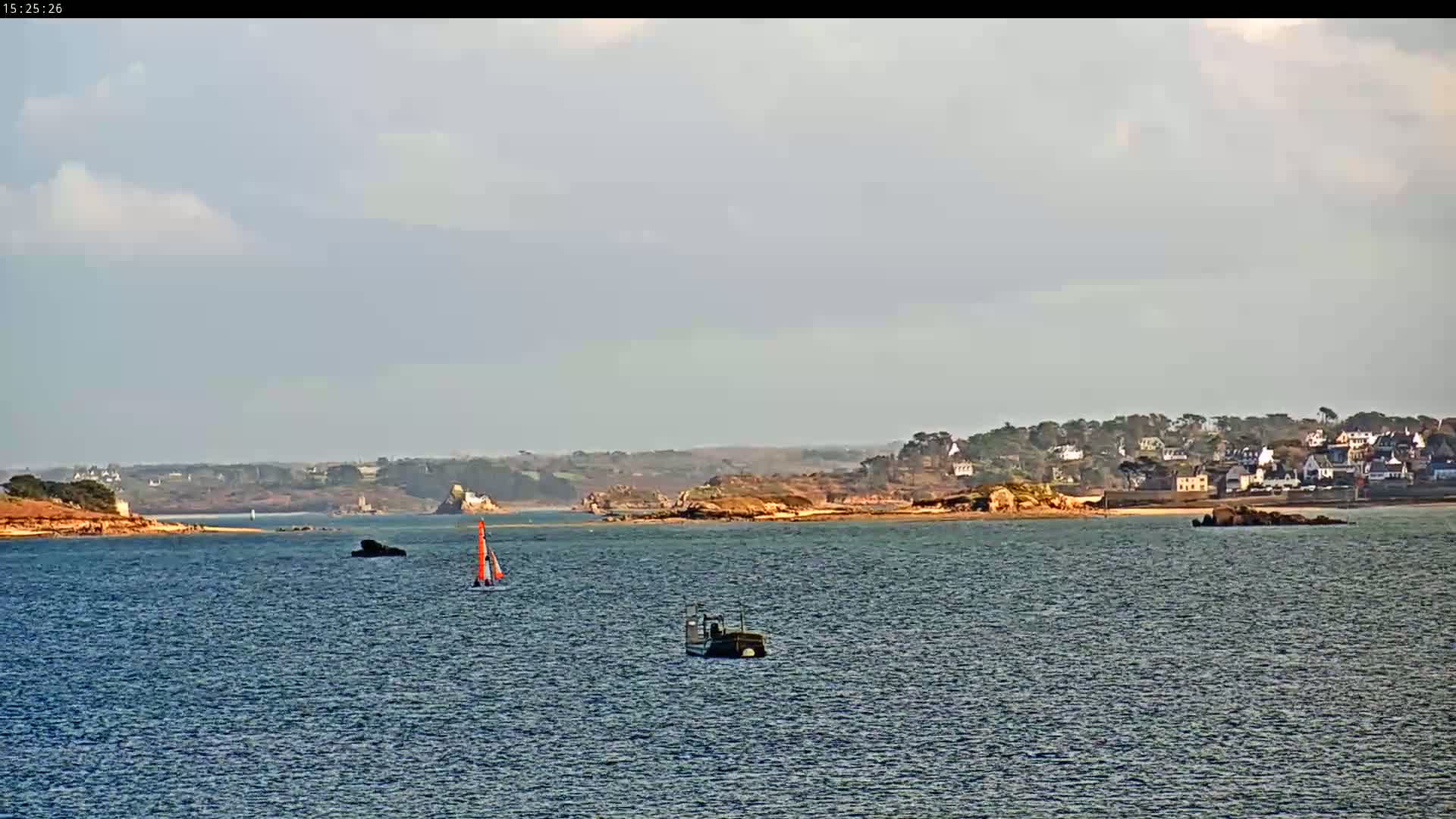 A sailboat and a small vessel are on a calm body of water in front of a distant coastline with a small town under a partly cloudy sky.