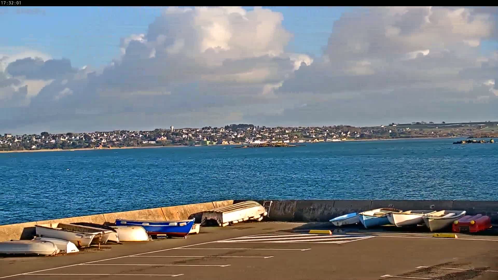 Several small boats are stored upside down and sideways on a paved area next to a body of water under a partly cloudy sky, with a town visible in the distance.