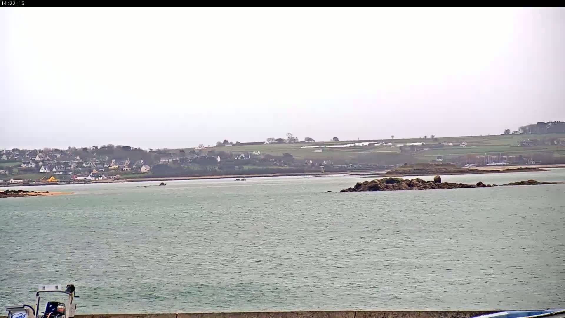 A body of water shows a small rocky island in the mid-ground, with a town and fields visible on the horizon under an overcast sky.