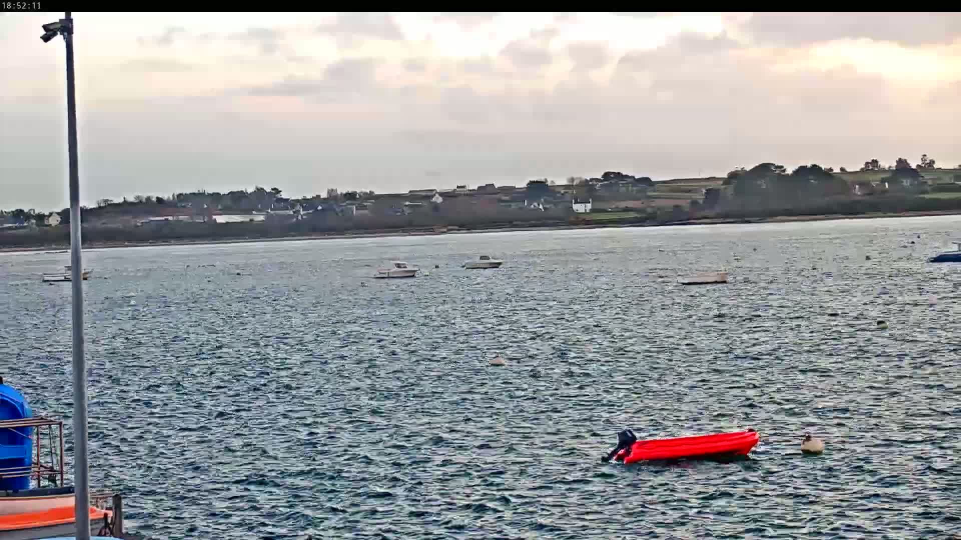 A mostly cloudy day shows a body of water with several small boats, a red inflatable boat, and a distant shoreline with buildings.
