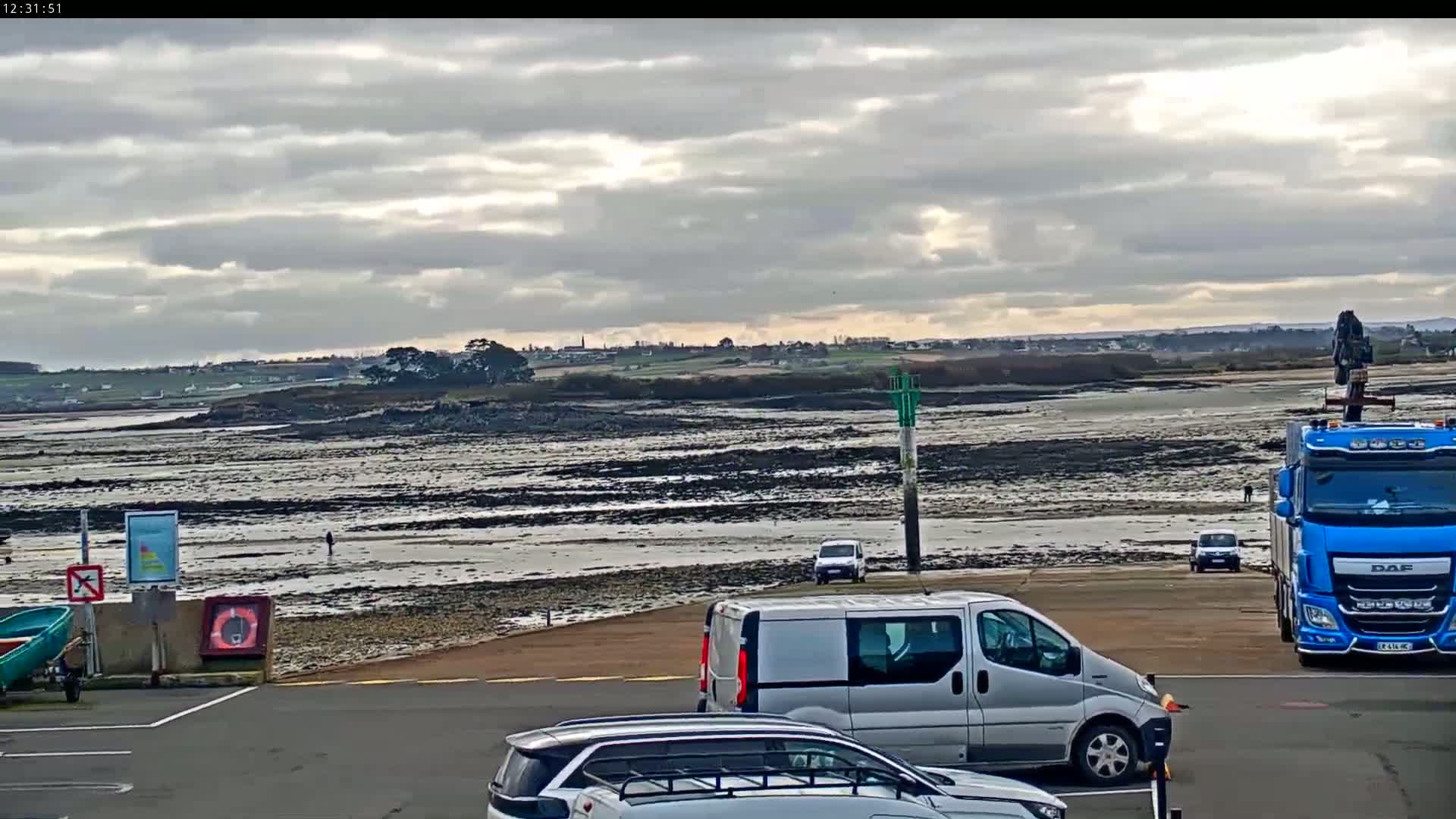 A mostly cloudy day shows a large expanse of exposed mudflats next to a parking lot with several vehicles, including a van and a large blue truck with a crane.