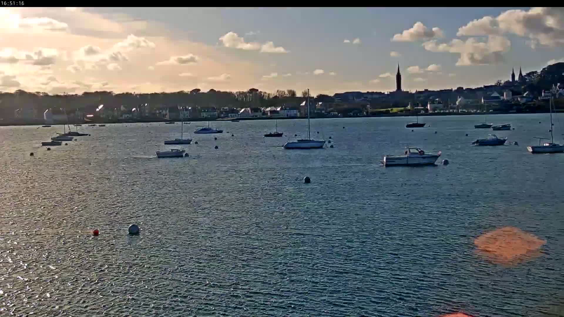 Many sailboats and motorboats are moored in a calm body of water under a partly cloudy sky, with a town visible in the background.