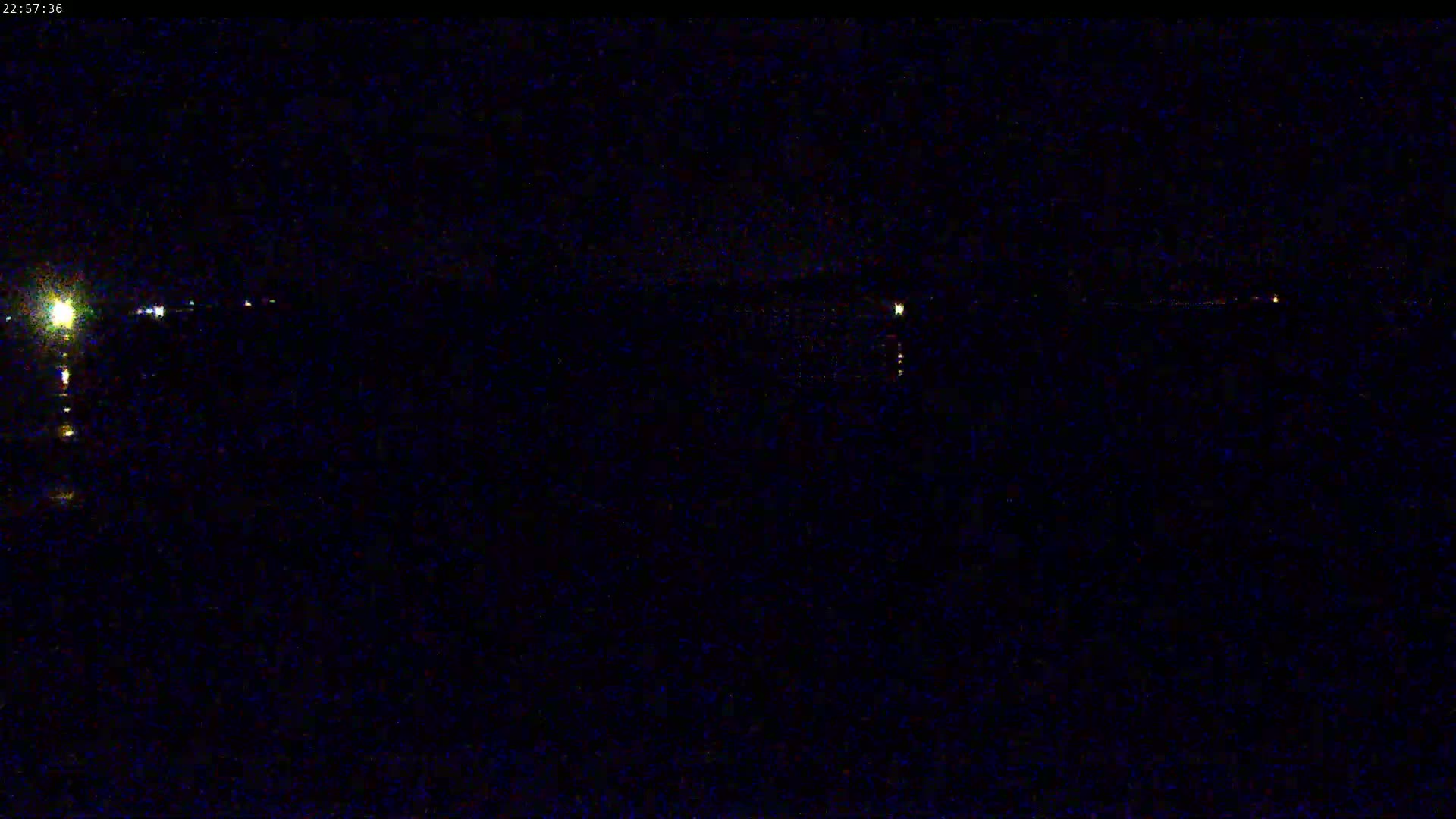 A nighttime view shows a dark body of water with a few distant, faint lights along the shoreline under a dark sky.