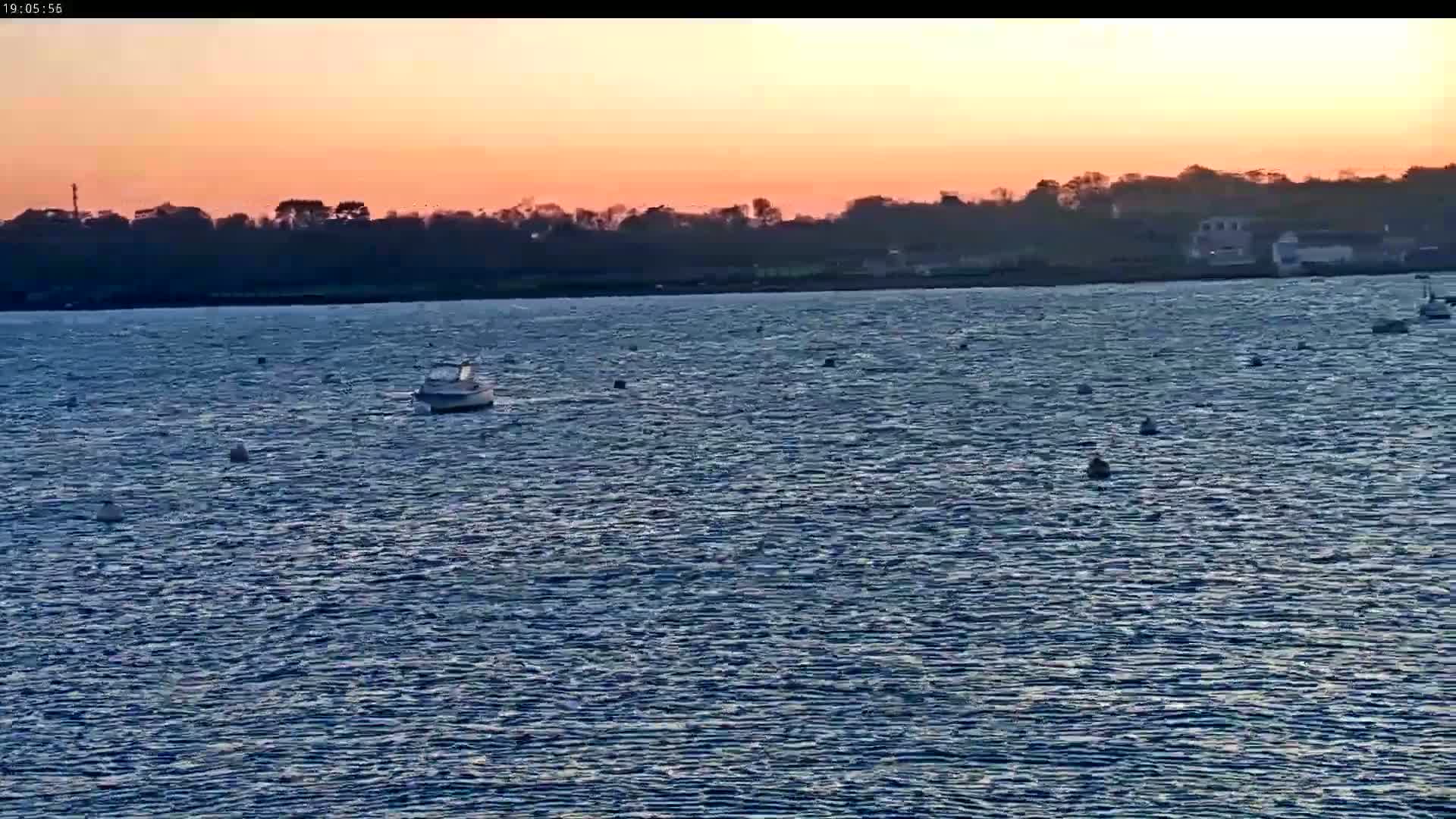 A small boat floats on a body of water at sunset under a mostly clear, pinkish-orange sky, with a dark shoreline visible in the distance.