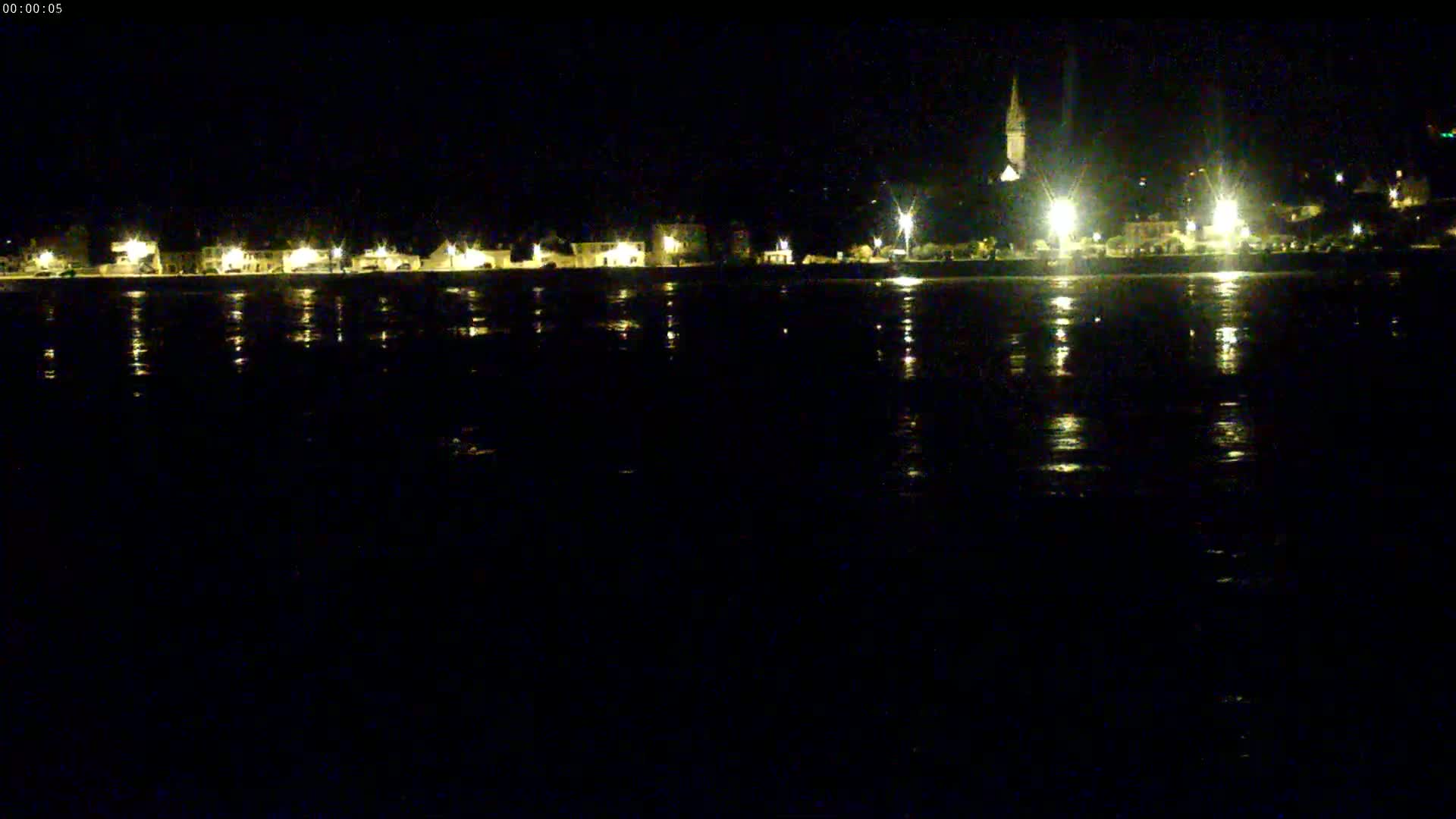 At night, a town's lights reflect on calm water, with a tall structure, possibly a church steeple, visible in the distance.