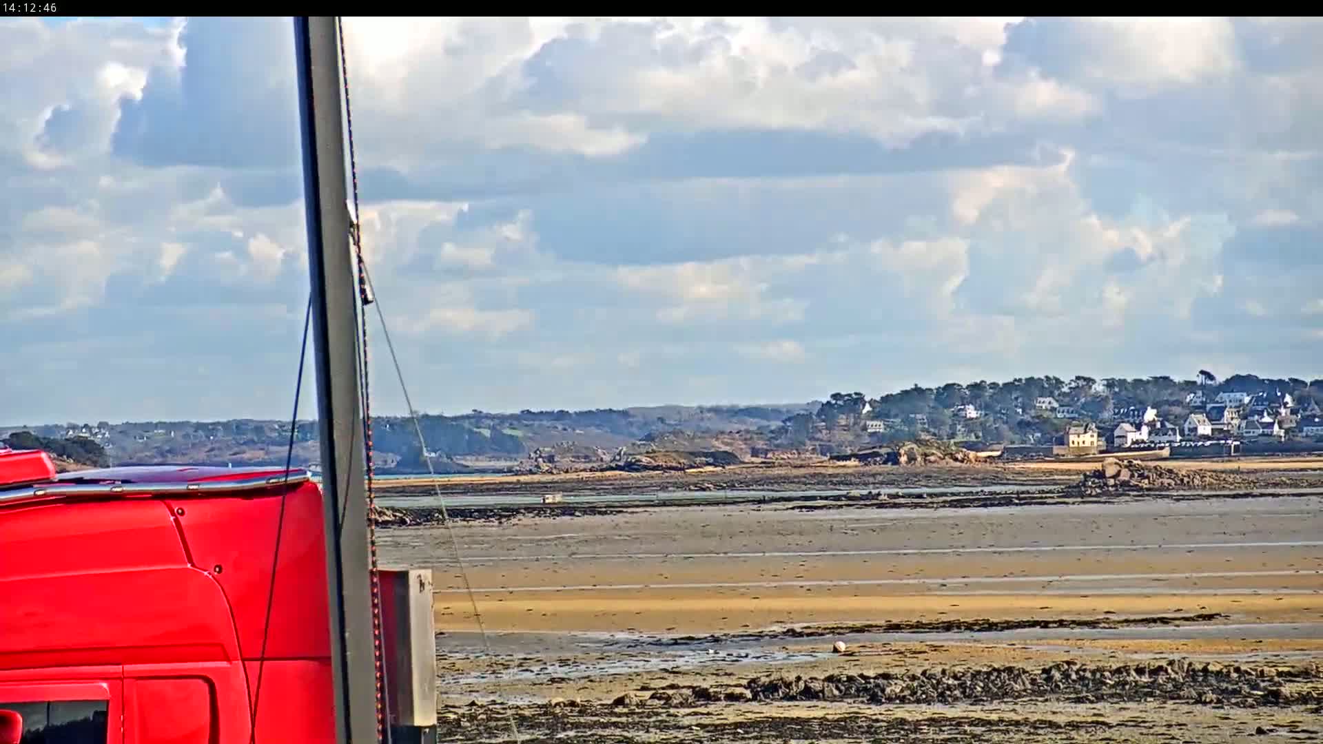 A partially visible red truck is parked near a wide expanse of exposed tidal flats under a partly cloudy sky, with a distant village visible across the water.