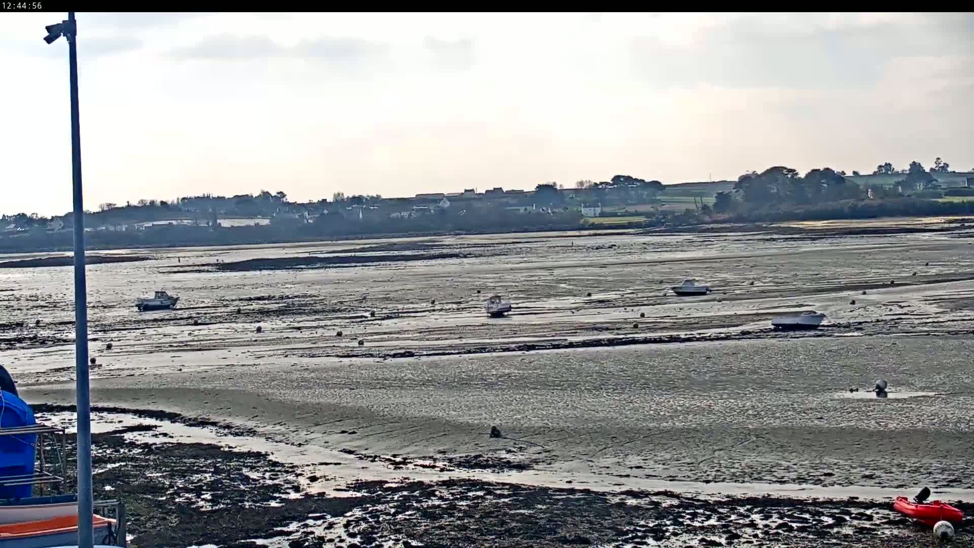 A wide expanse of mudflats at low tide is dotted with several small boats, under an overcast sky, with a distant shoreline visible.