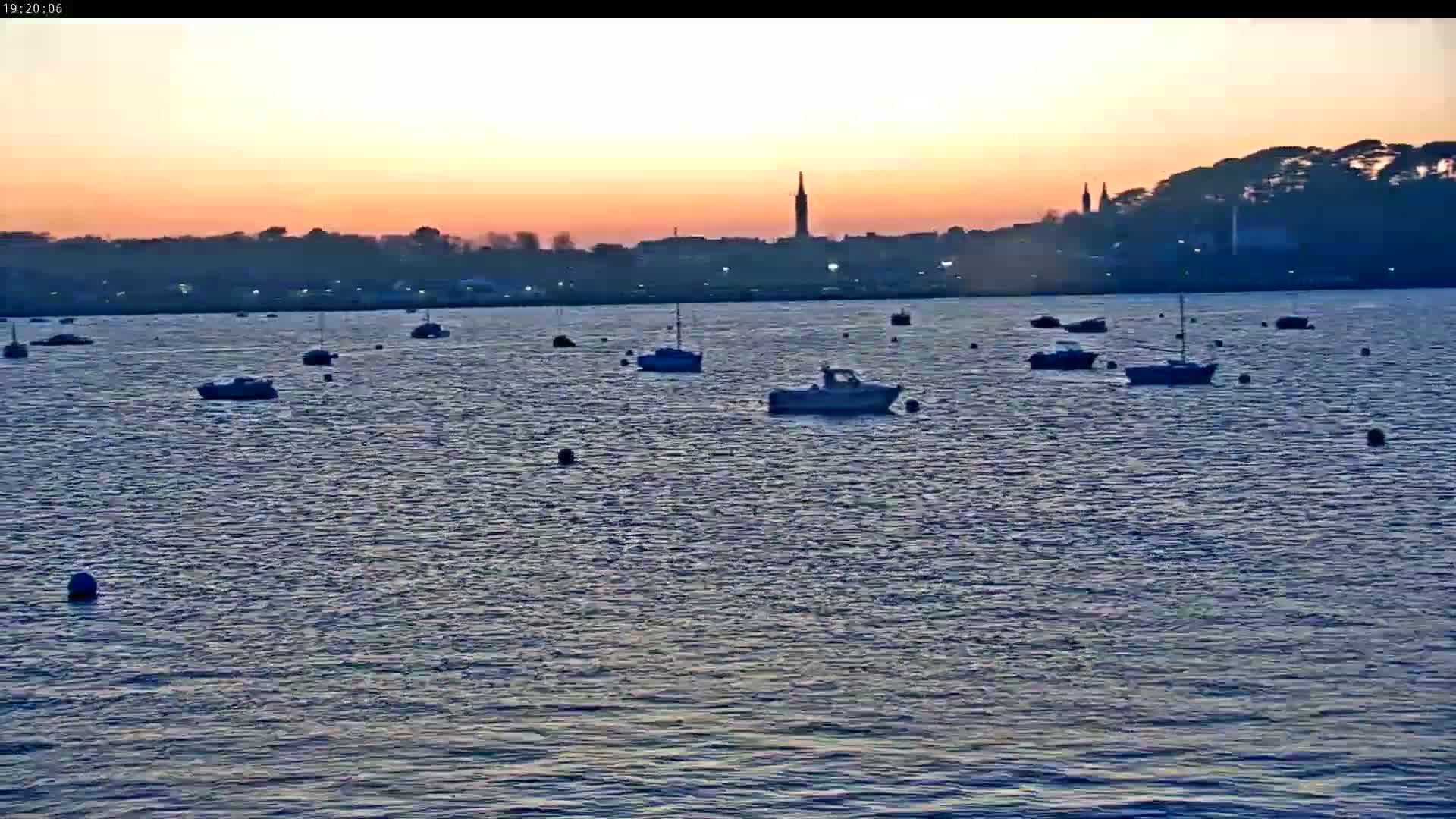 Several boats are moored on calm water at sunset, with a town silhouetted on the horizon.