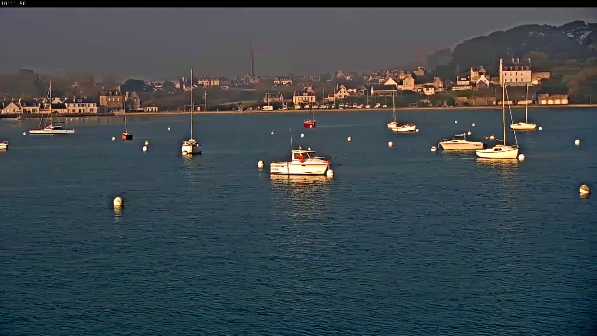 Several boats are moored in a calm body of water in front of a hazy coastal town.