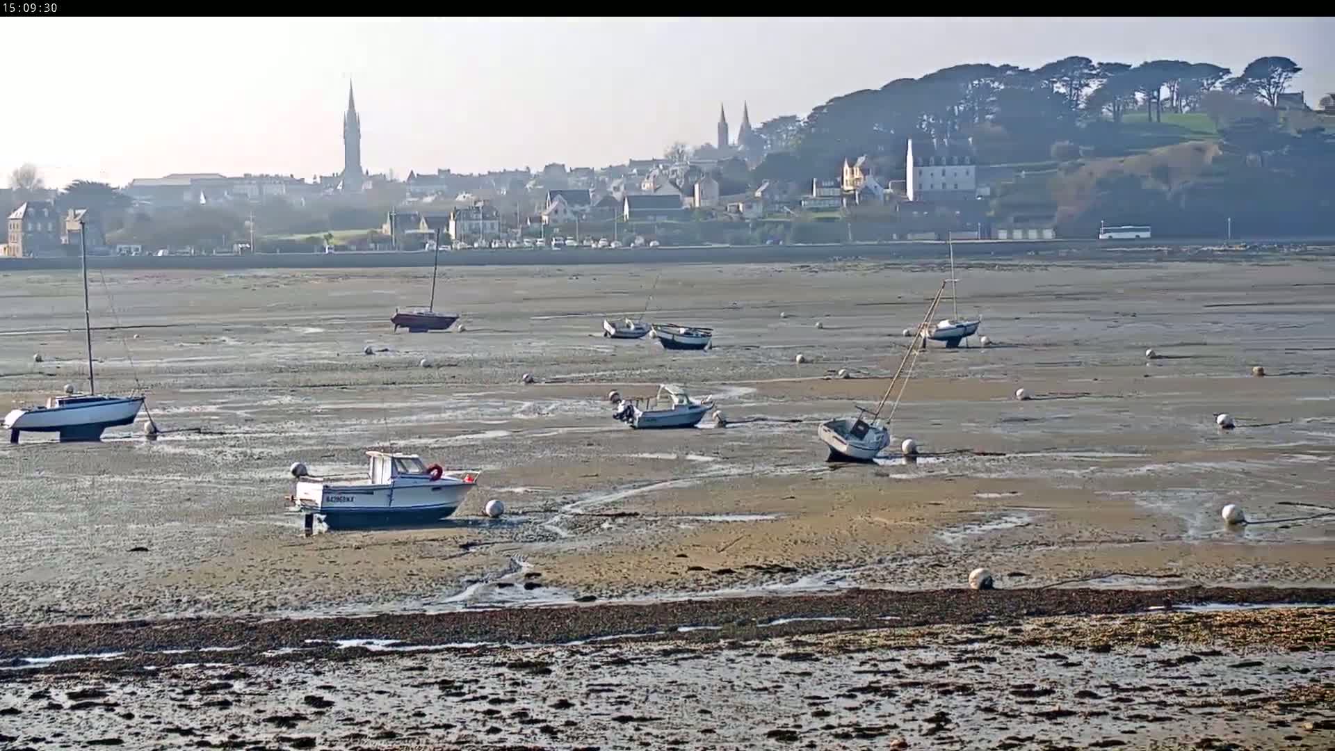 Several small boats sit on a mostly exposed, sandy seabed near a distant town under a hazy, sunny sky.