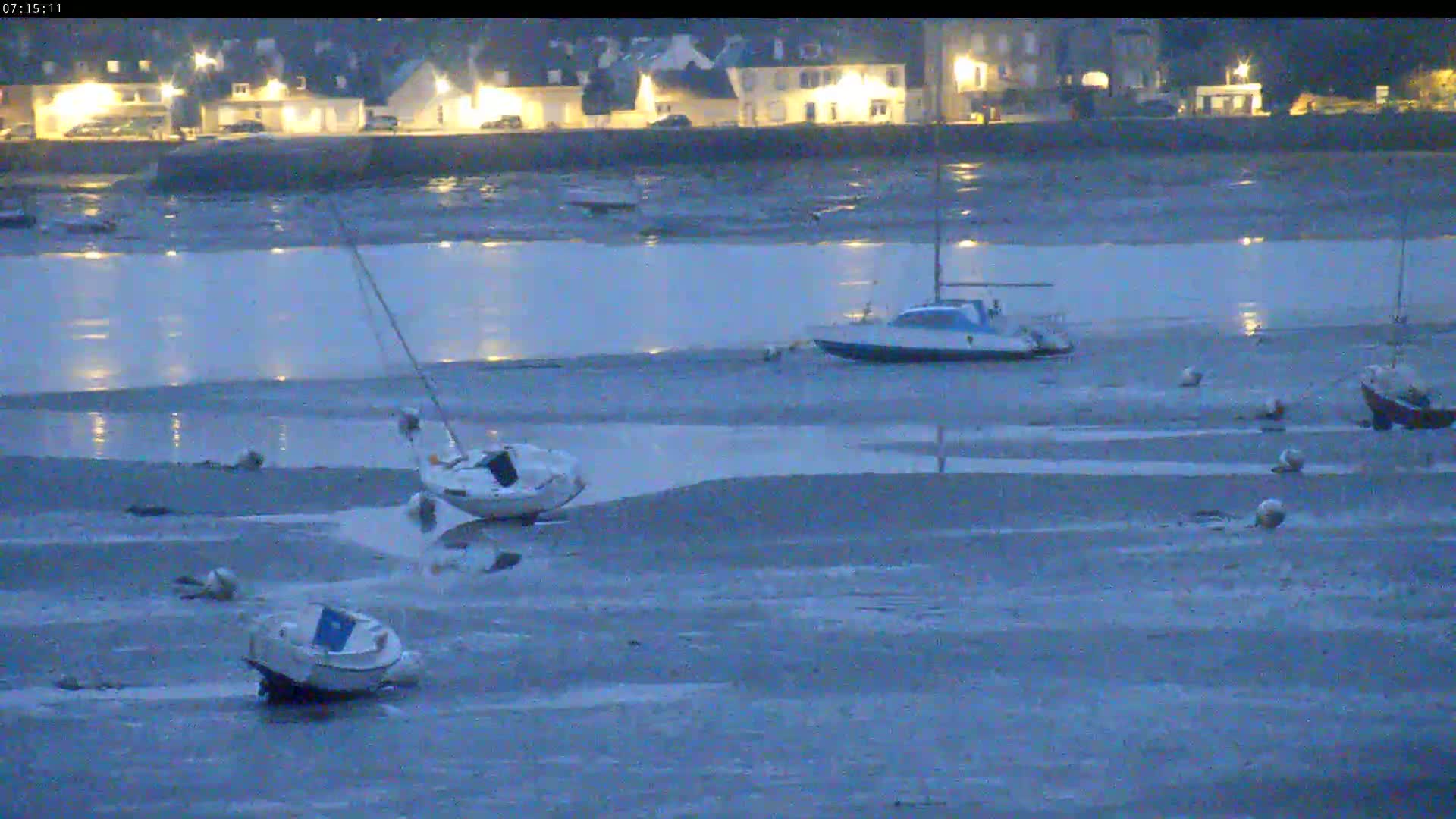 Several small boats are beached on a mudflat at low tide in front of a dimly lit waterfront village at dusk.