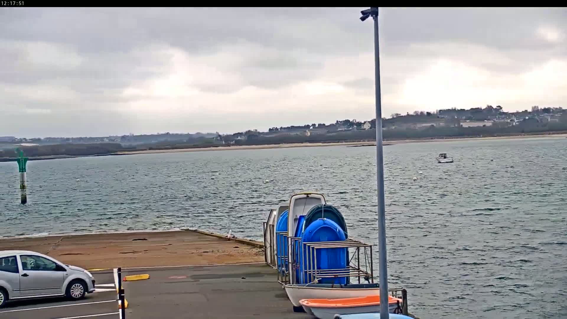A silver car is parked near a rack of blue and white boats on a dock overlooking a choppy body of water under a cloudy sky.
