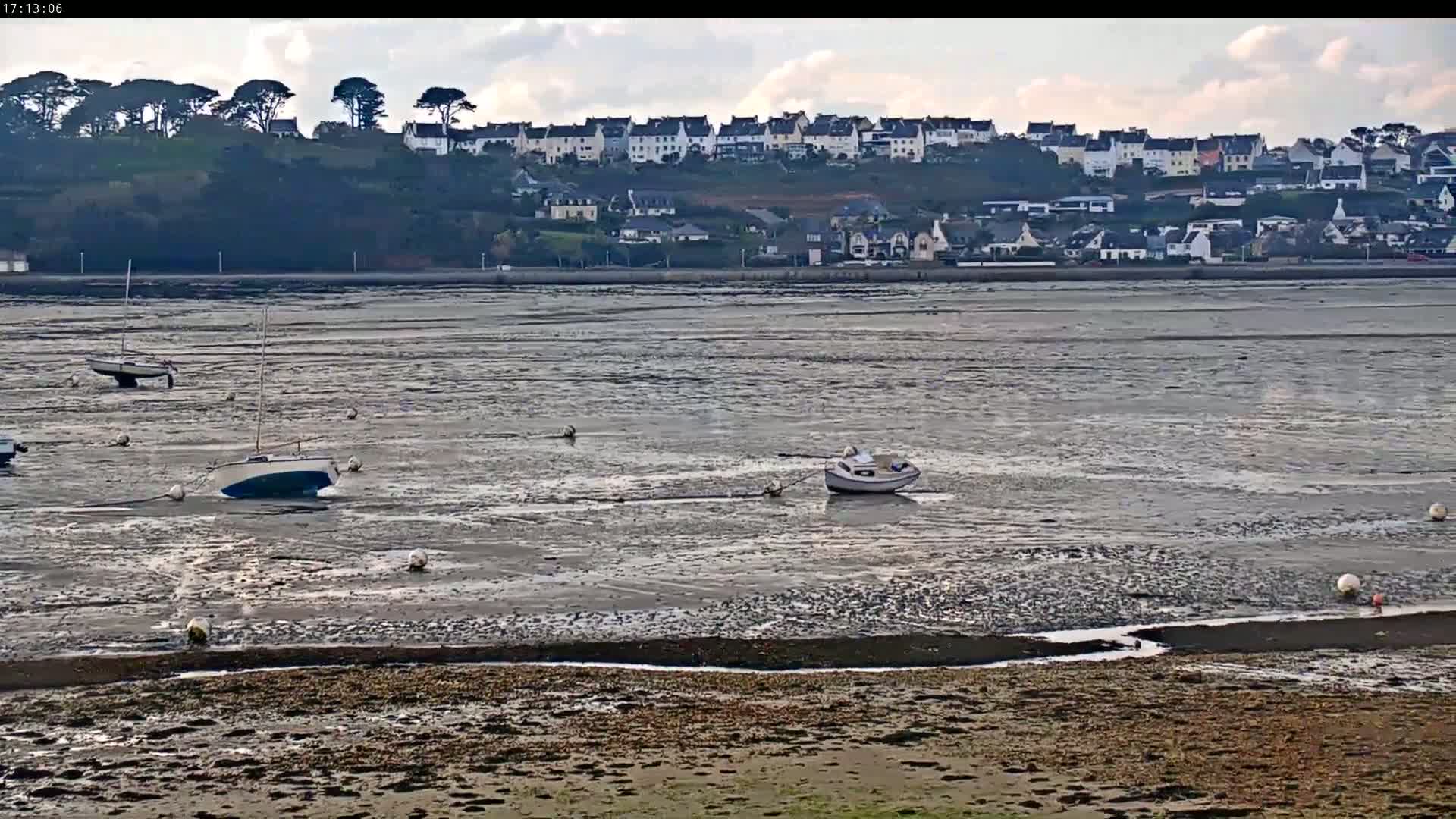 Several small boats are grounded in the mudflats of a bay, with a village visible on the far shore under a partly cloudy sky.