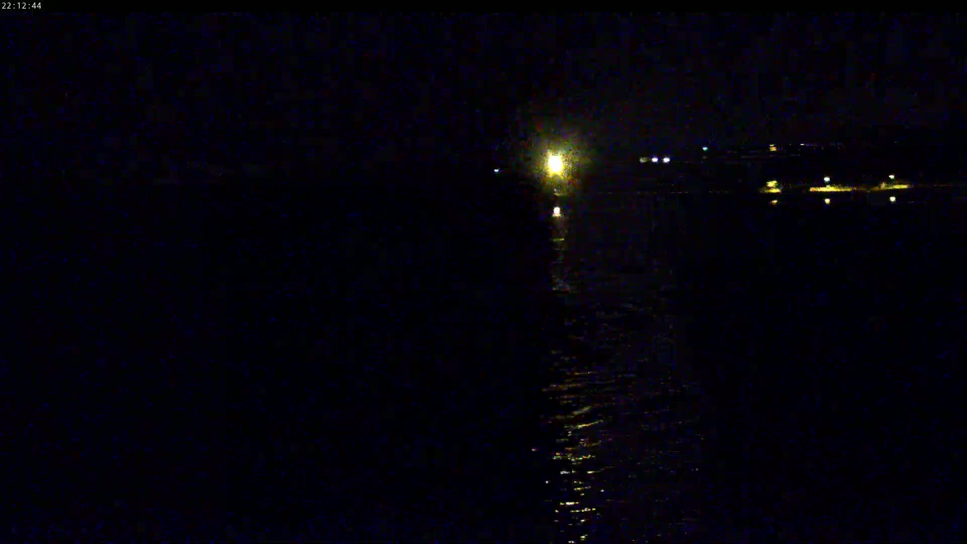 A dark, nighttime view of a body of water with faint lights on the far shore and a brighter light source reflecting on the water in the center.