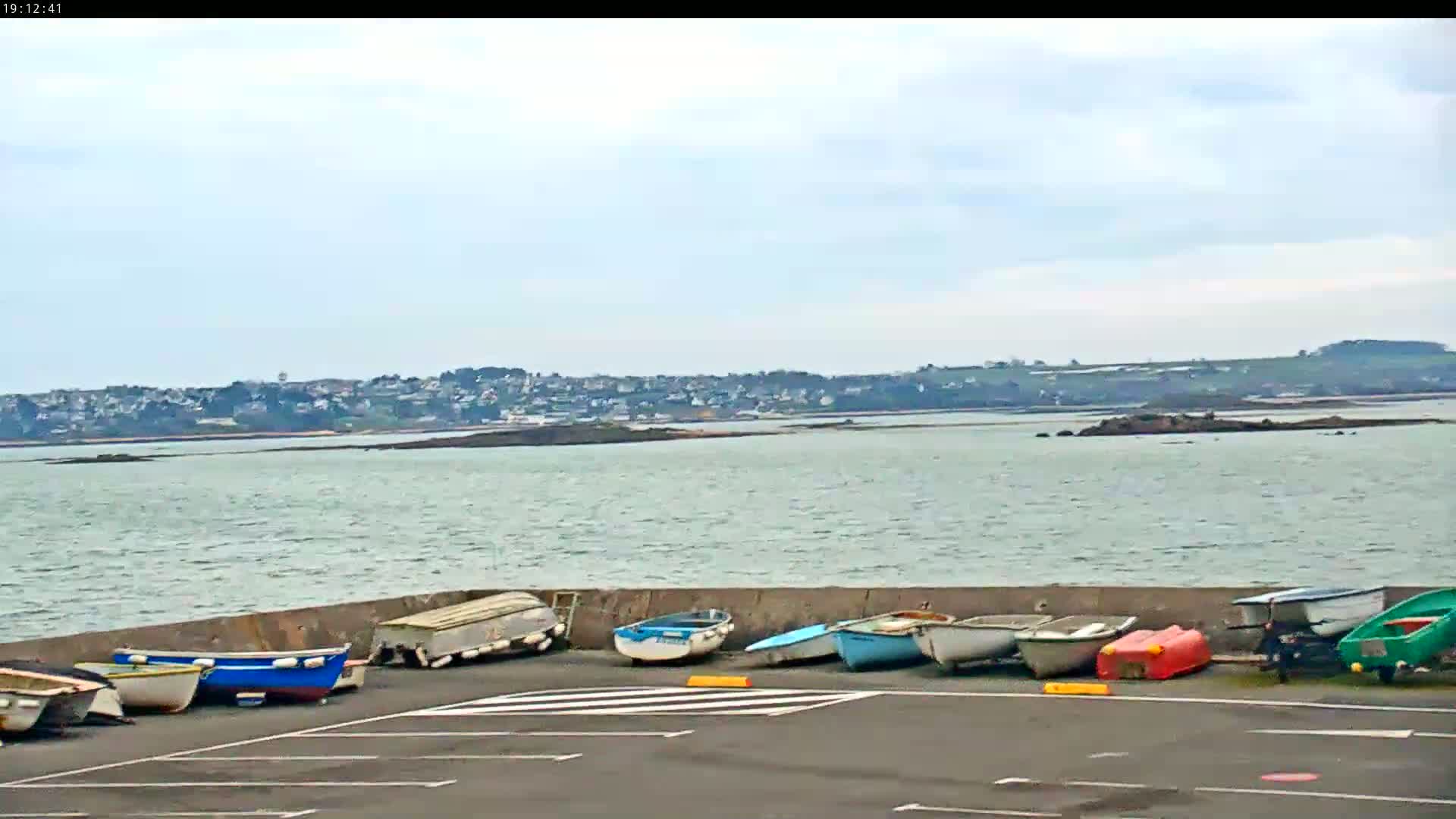 Several small boats are parked on a paved area next to a calm body of water under a mostly cloudy sky, with a distant town visible across the water.