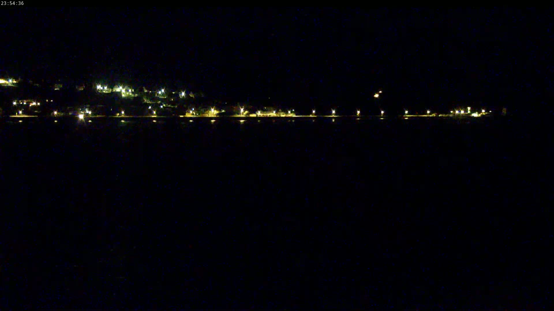 A nighttime view of a dark body of water with a line of lights on the far shore illuminating a hilly area.