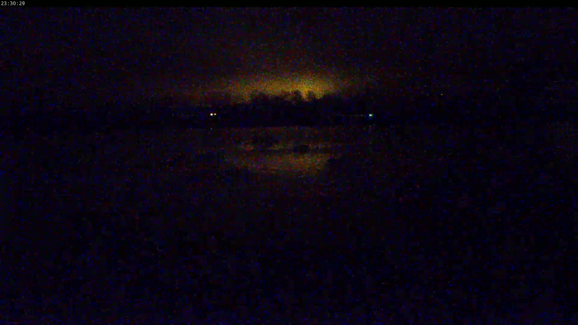 A dark, nighttime scene shows a dimly lit shoreline with trees and water reflecting a faint, yellowish glow in the distance under an overcast sky.