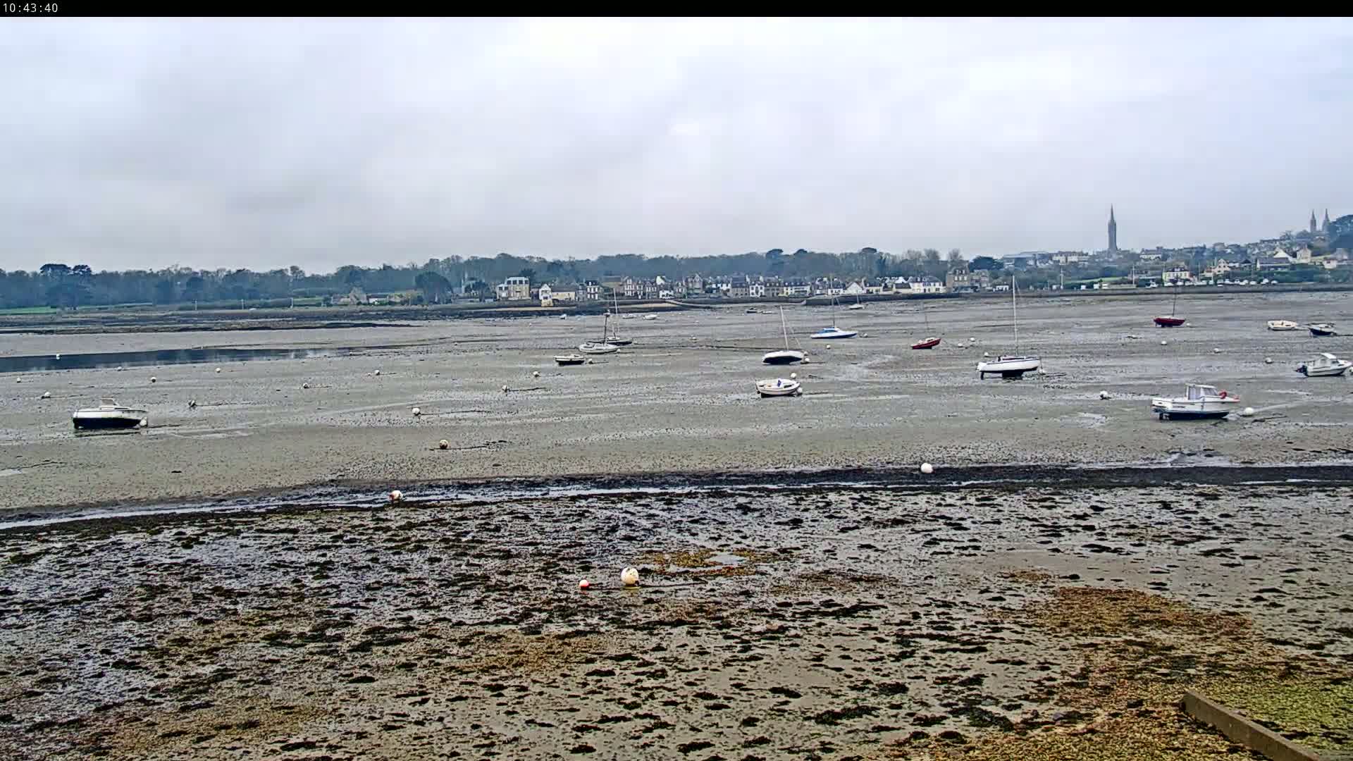 A wide expanse of mudflats at low tide reveals numerous small boats resting in the shallow water, with a distant town visible under a cloudy sky.