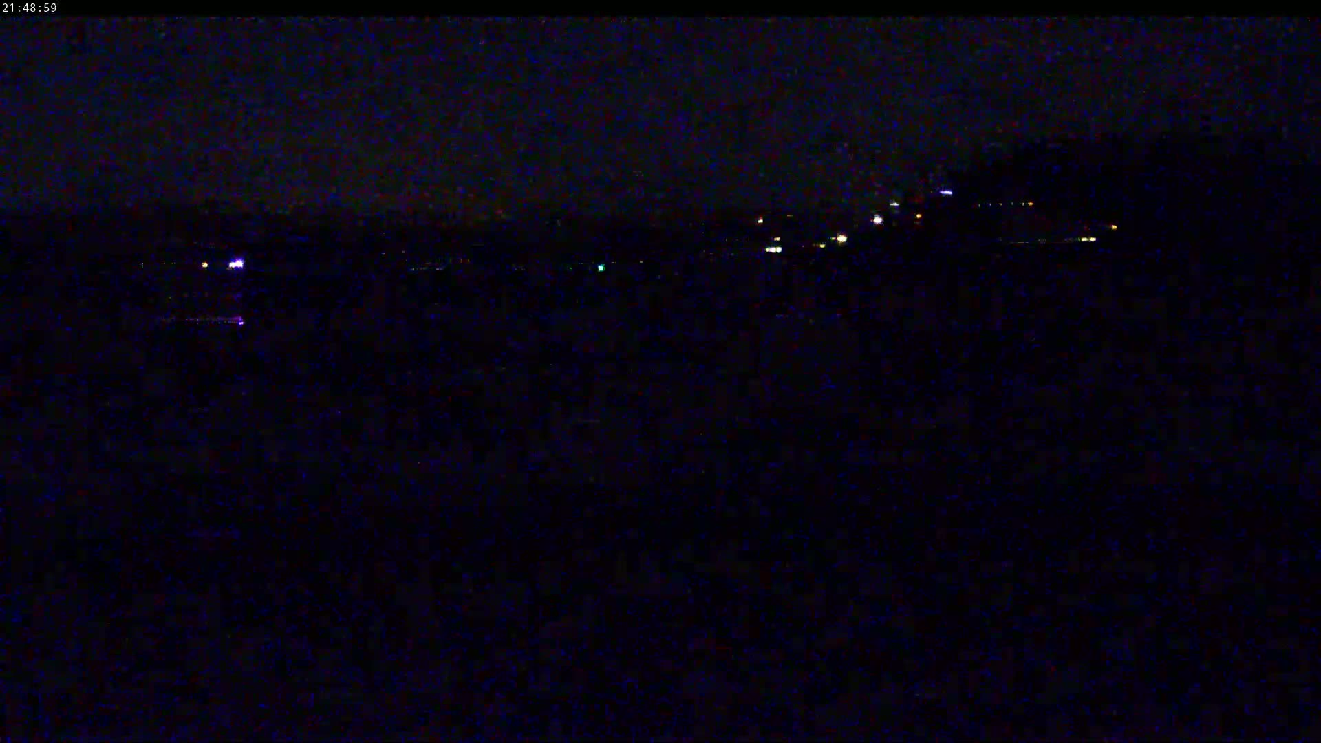 A dark nighttime scene shows a distant line of lights across a dark body of water under a clear, starless sky.
