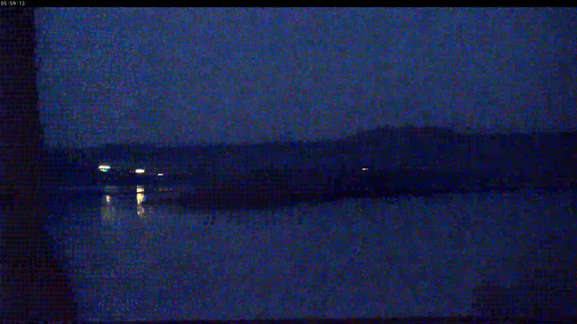 A dark, nighttime view shows a body of still water reflecting a few distant lights from the opposite shore, under a clear, dark sky.