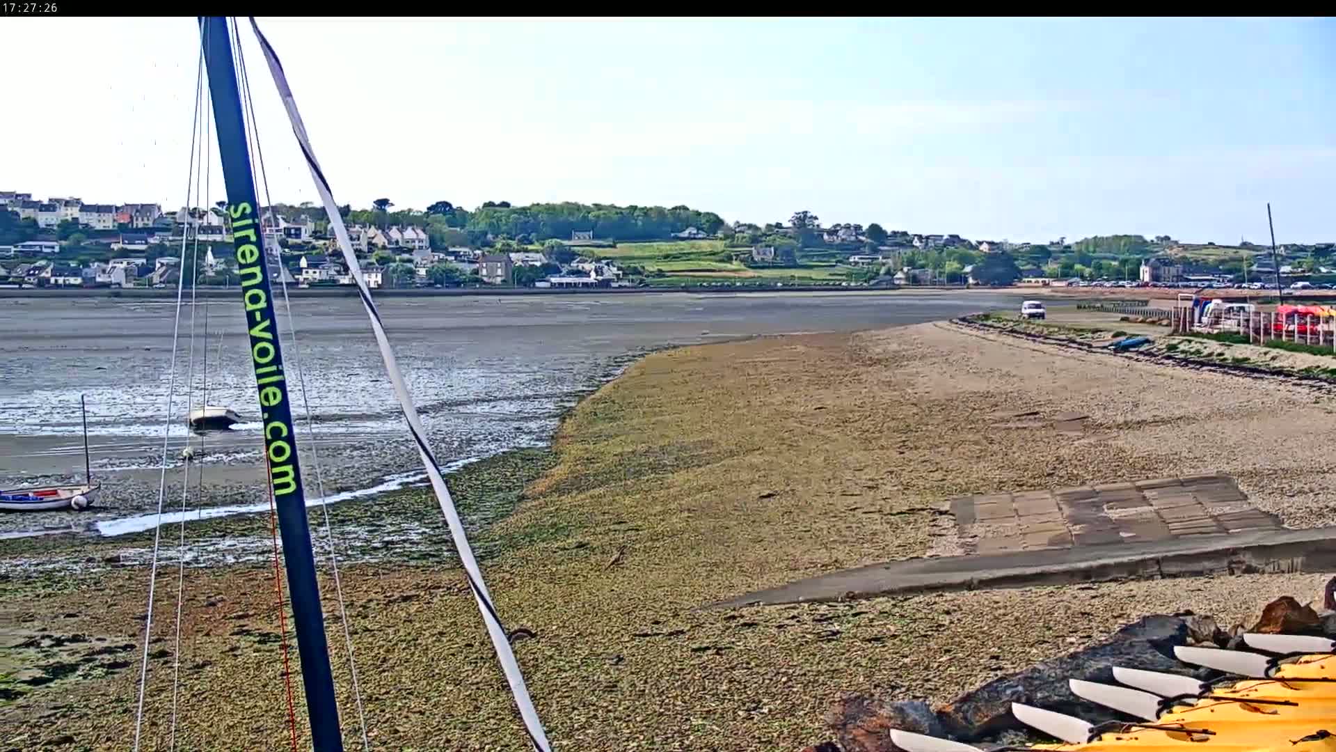 A sandy beach with several kayaks and a sailboat mast in the foreground, extends to a calm body of water, with a town visible in the background under a mostly clear sky.