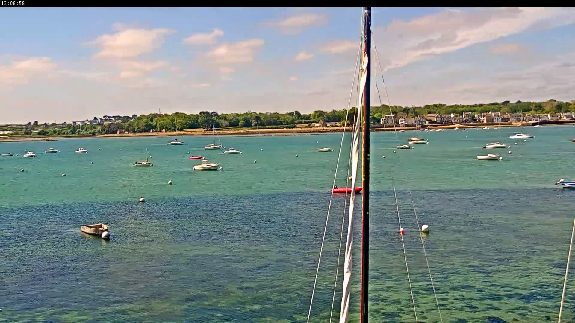 A calm, sunny day shows a clear, teal-colored body of water with numerous sailboats and small motorboats scattered across the surface, a shoreline with green trees and buildings visible in the distance.