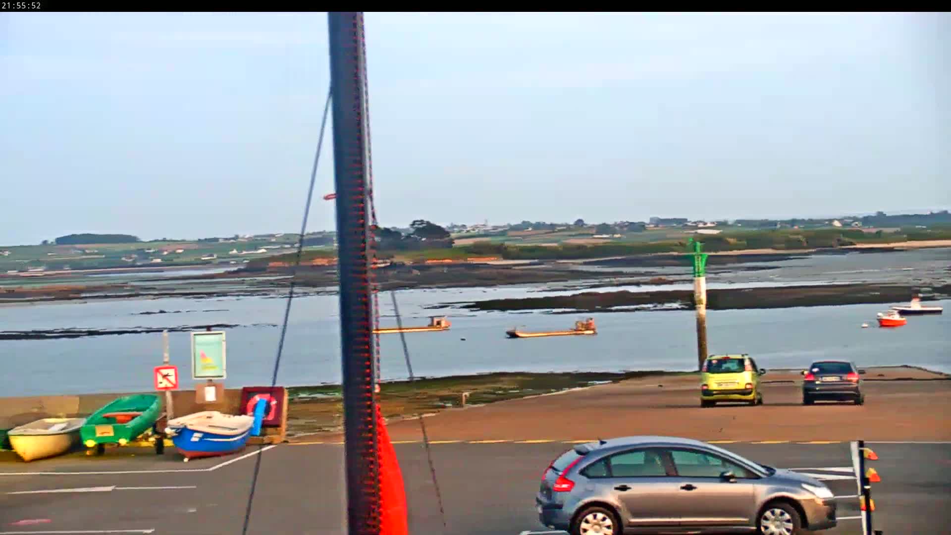 A calm body of water with several small boats, cars parked in a lot, and a distant shoreline under a clear sky.