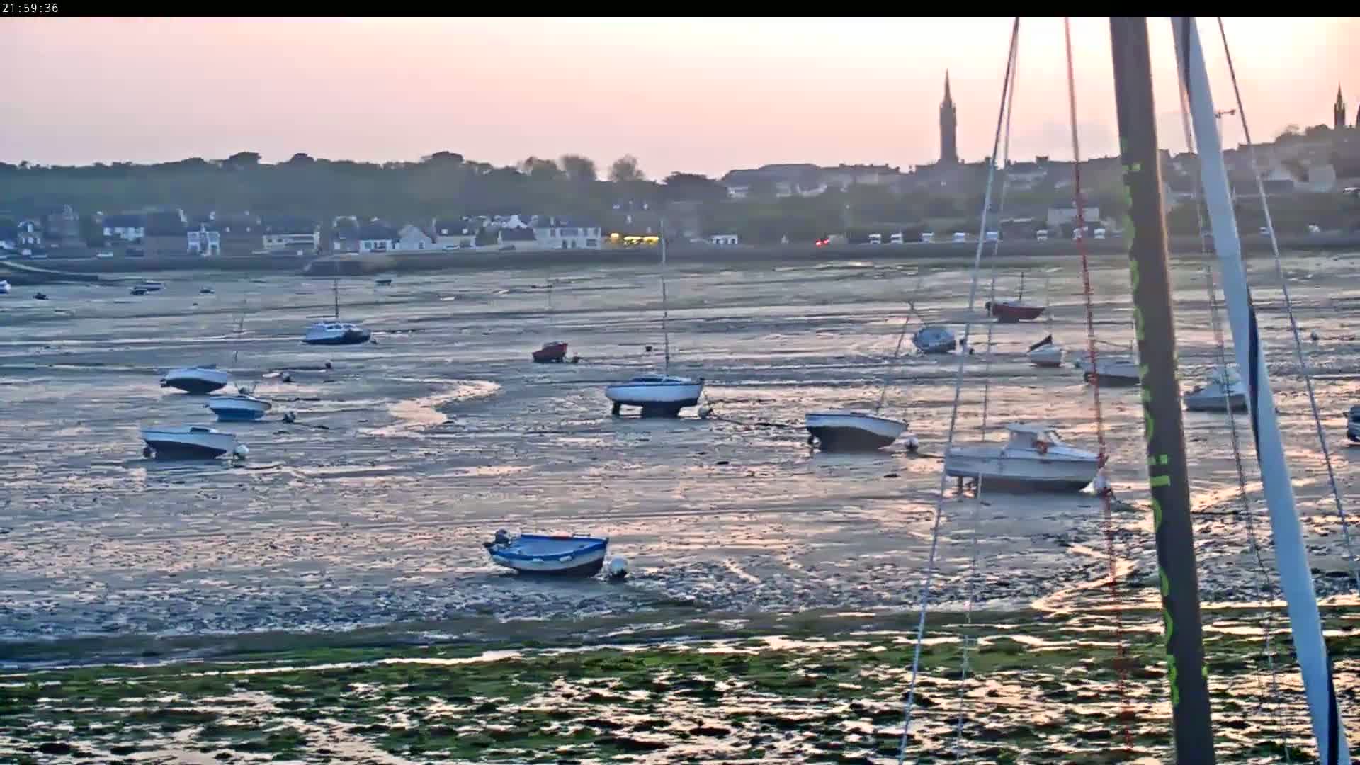 Many small boats sit on exposed mudflats at low tide in front of a town at sunset.