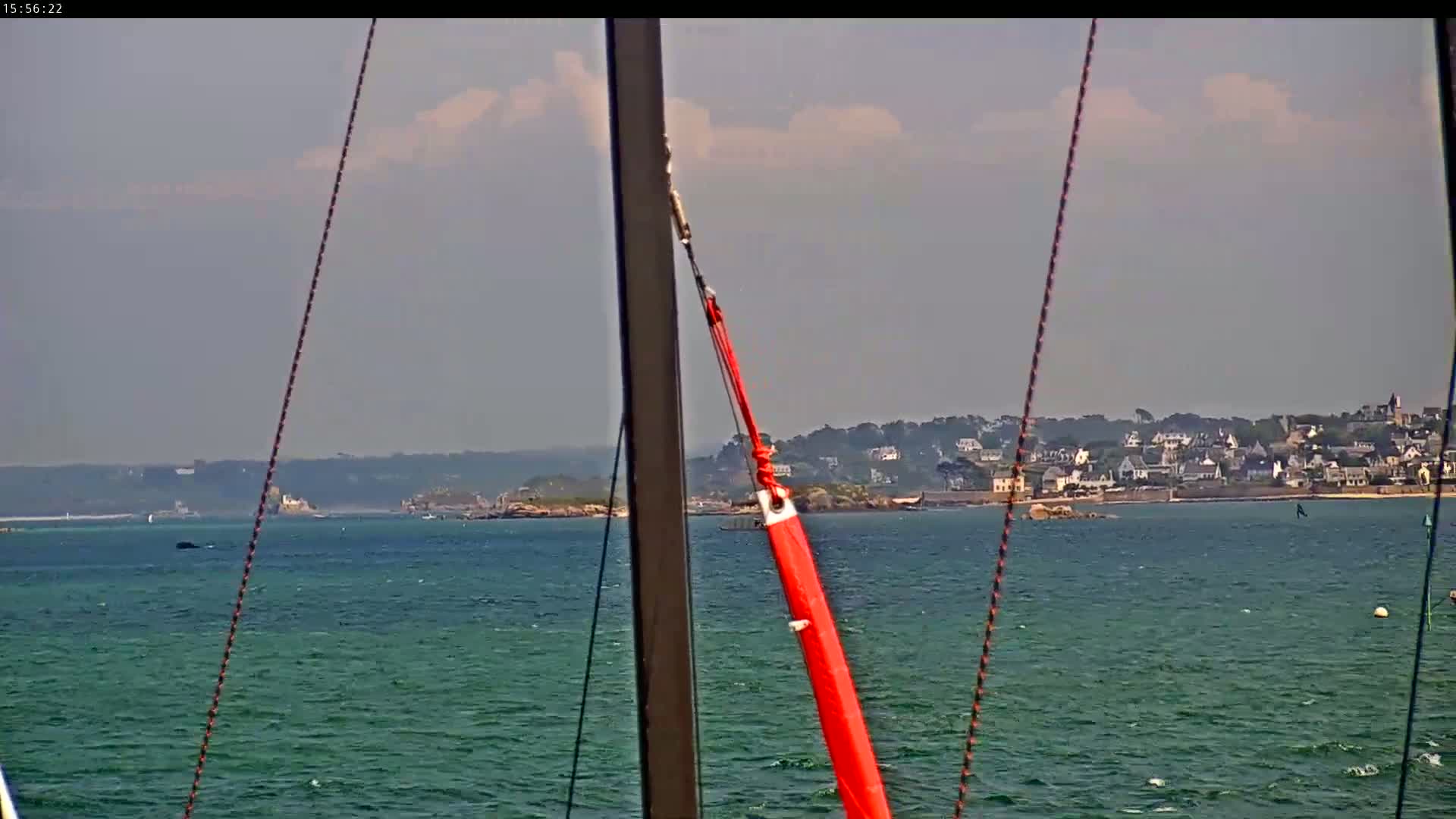 A mostly sunny day at sea shows a coastal town in the distance viewed from a sailboat's mast.
