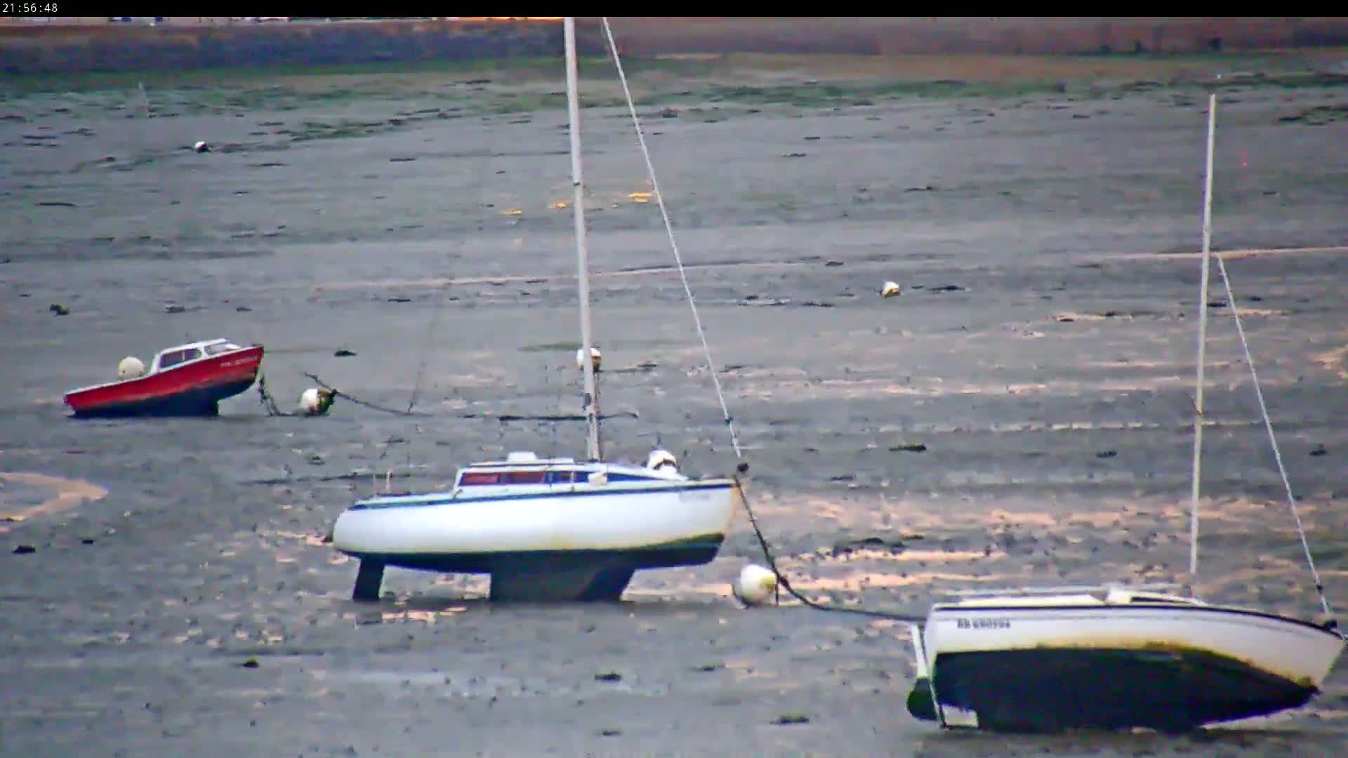 Three small boats, including two sailboats and a motorboat, are resting on a tidal mudflat under a calm sky.