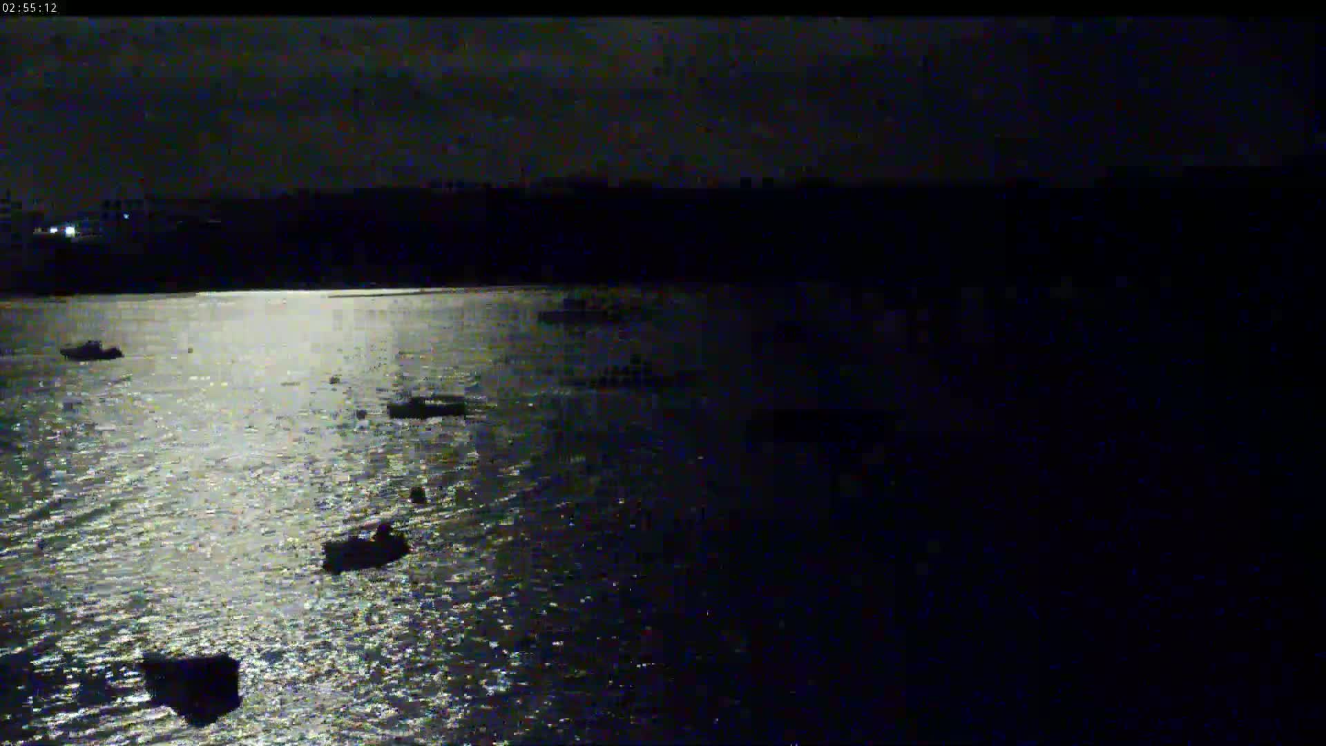 Several small boats are moored on a dark, moonlit body of water under a mostly cloudy night sky.