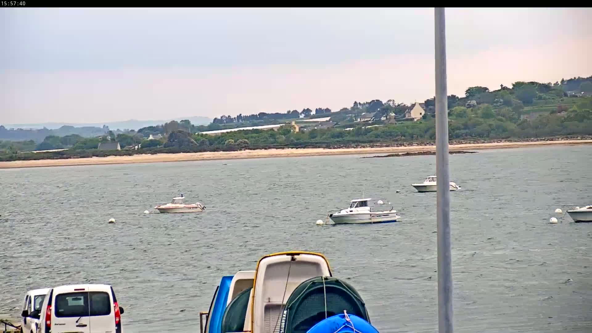 Several boats are moored in a calm, gray body of water, with a sandy beach and distant green hills visible under an overcast sky.