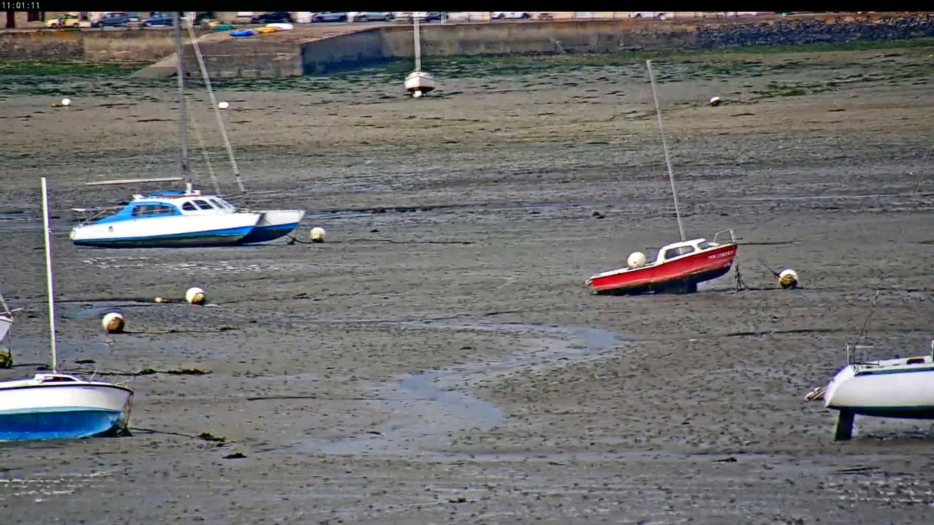 Several small boats sit on a mudflat at low tide under a bright, sunny sky.