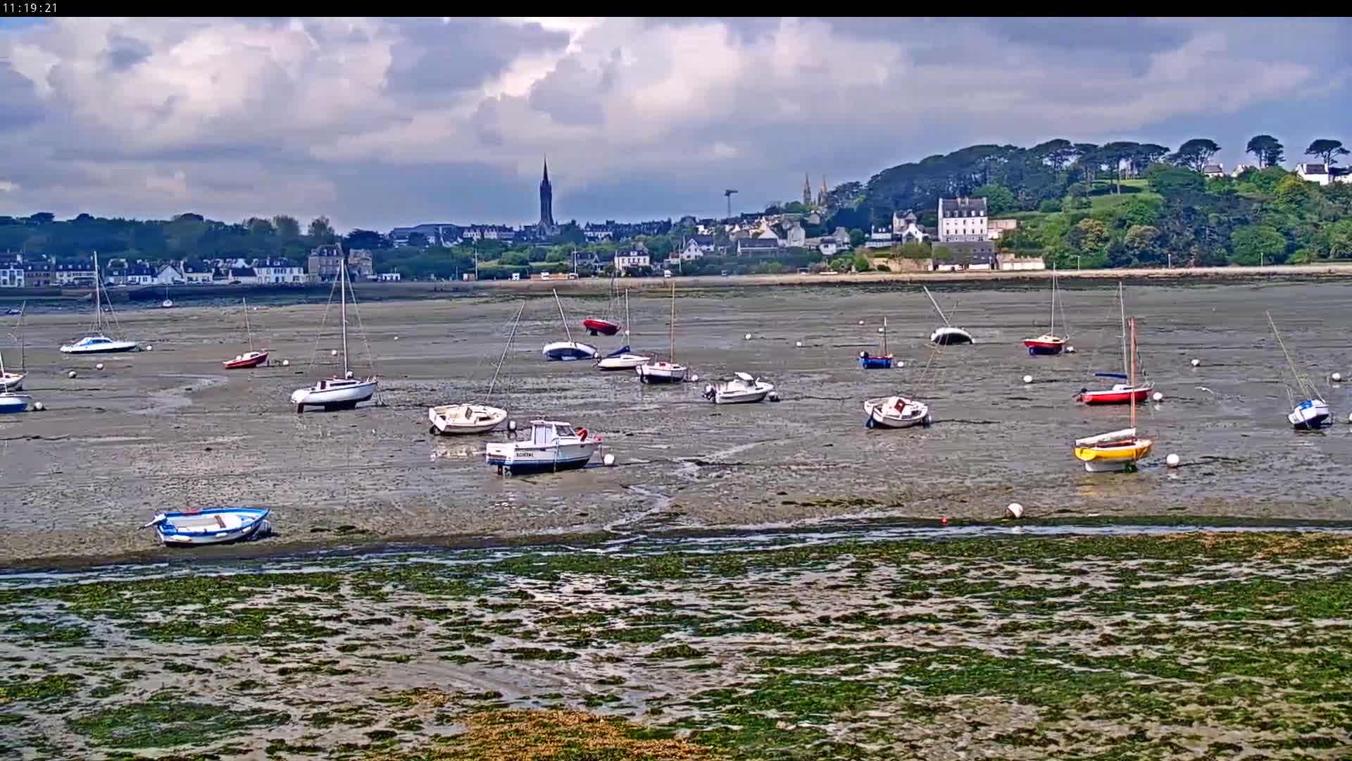 Numerous small sailboats and motorboats are resting on a wide expanse of exposed mudflats under a partly cloudy sky, with a distant town visible on the horizon.
