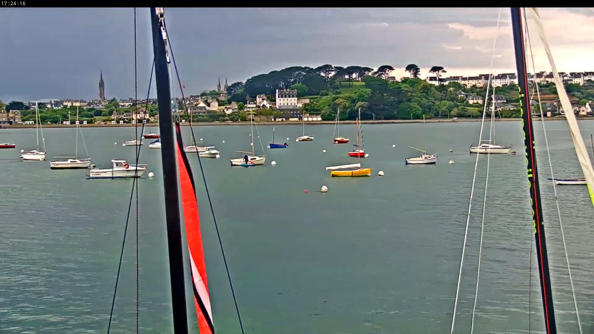 Numerous sailboats and motorboats are moored in calm water in front of a town nestled beside a verdant hill under a cloudy sky.