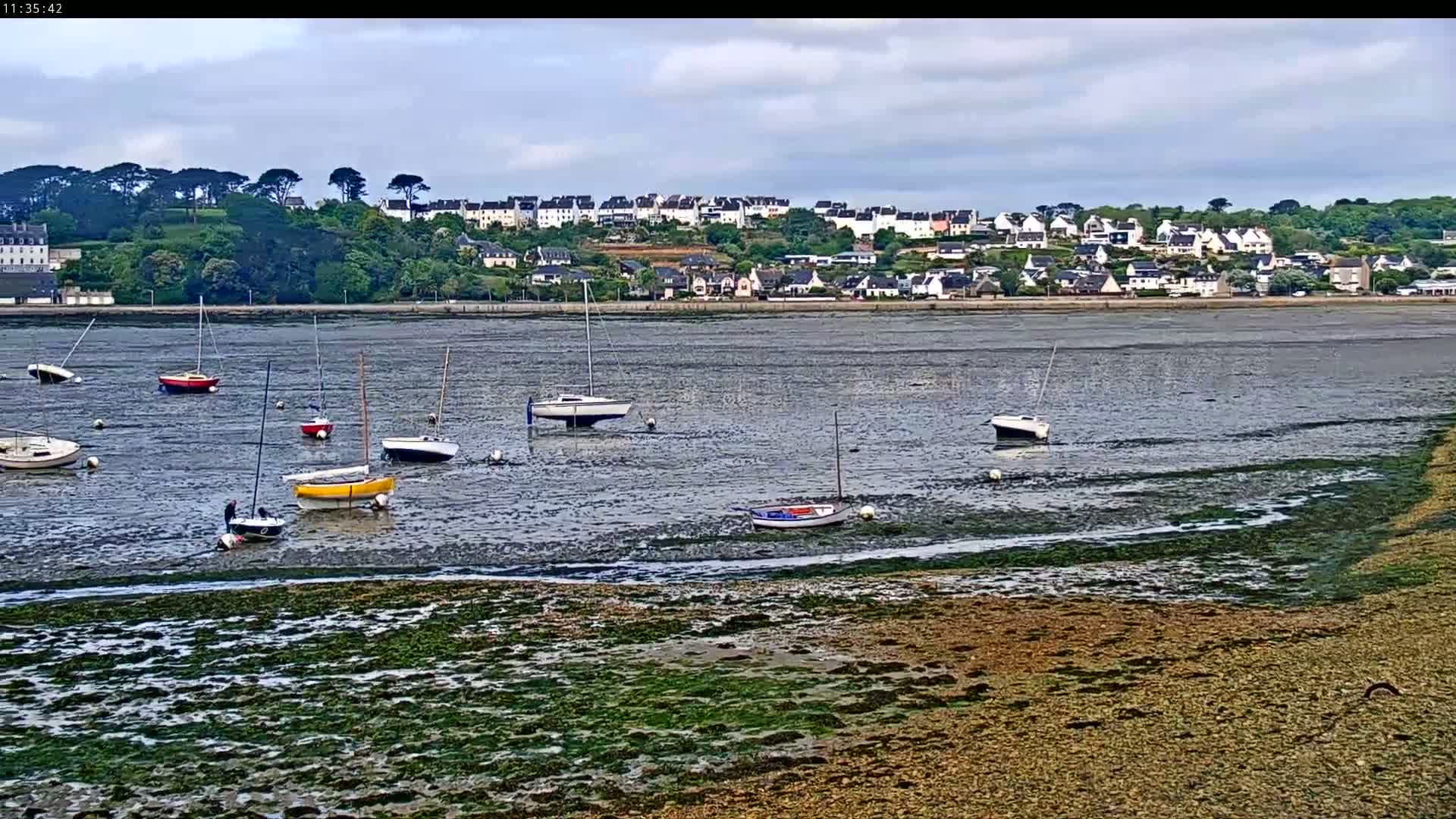 Several sailboats are moored in shallow, muddy water at low tide, with a village nestled on a hillside in the background under a cloudy sky.