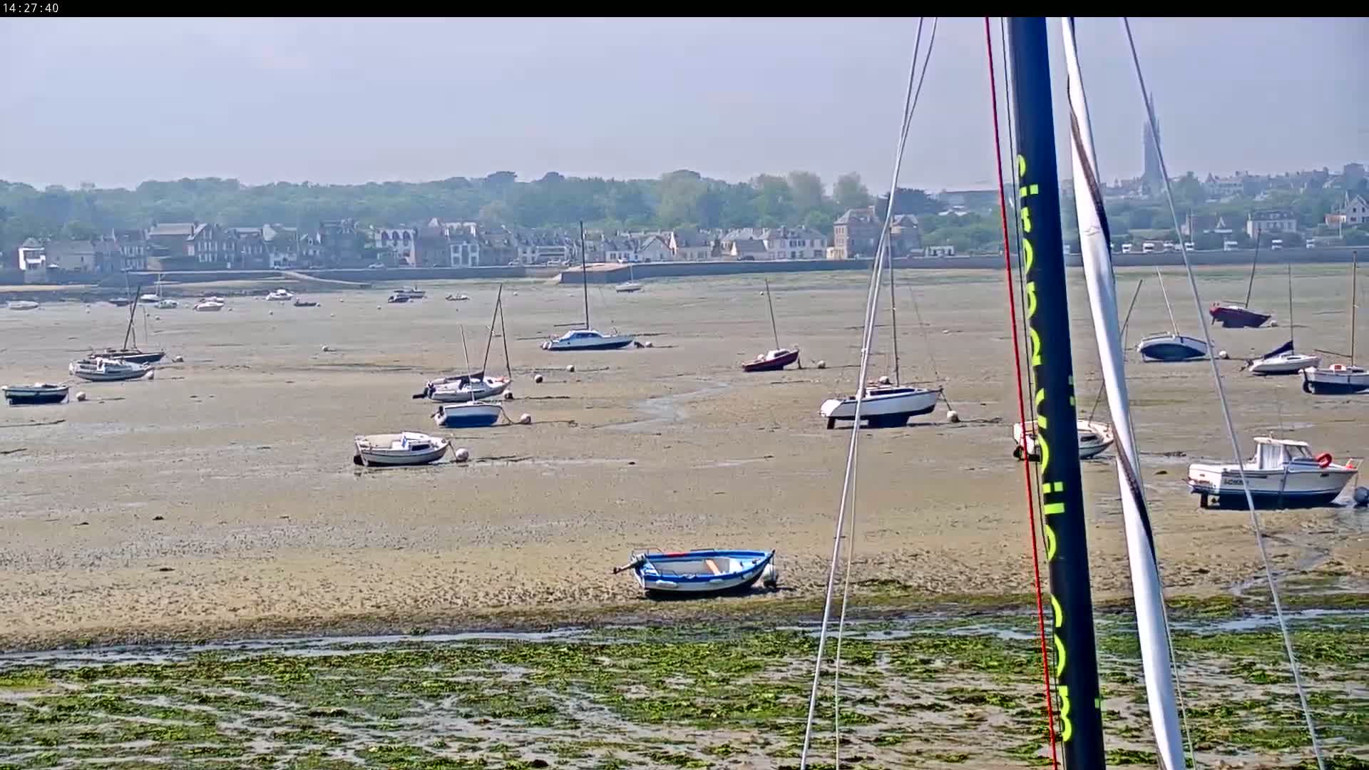 Many sailboats and small motorboats are beached on a wide, sandy expanse at low tide under a sunny sky, with a distant town visible in the background.