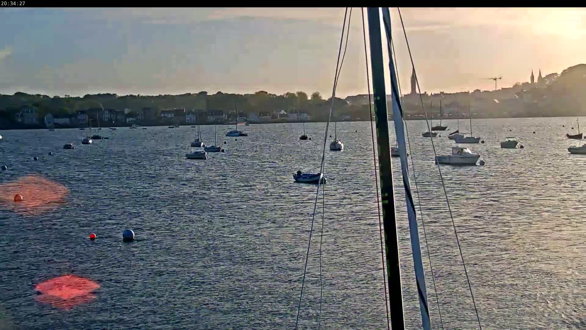 Numerous small boats are moored in calm water near a distant town under a partly cloudy sunset sky.