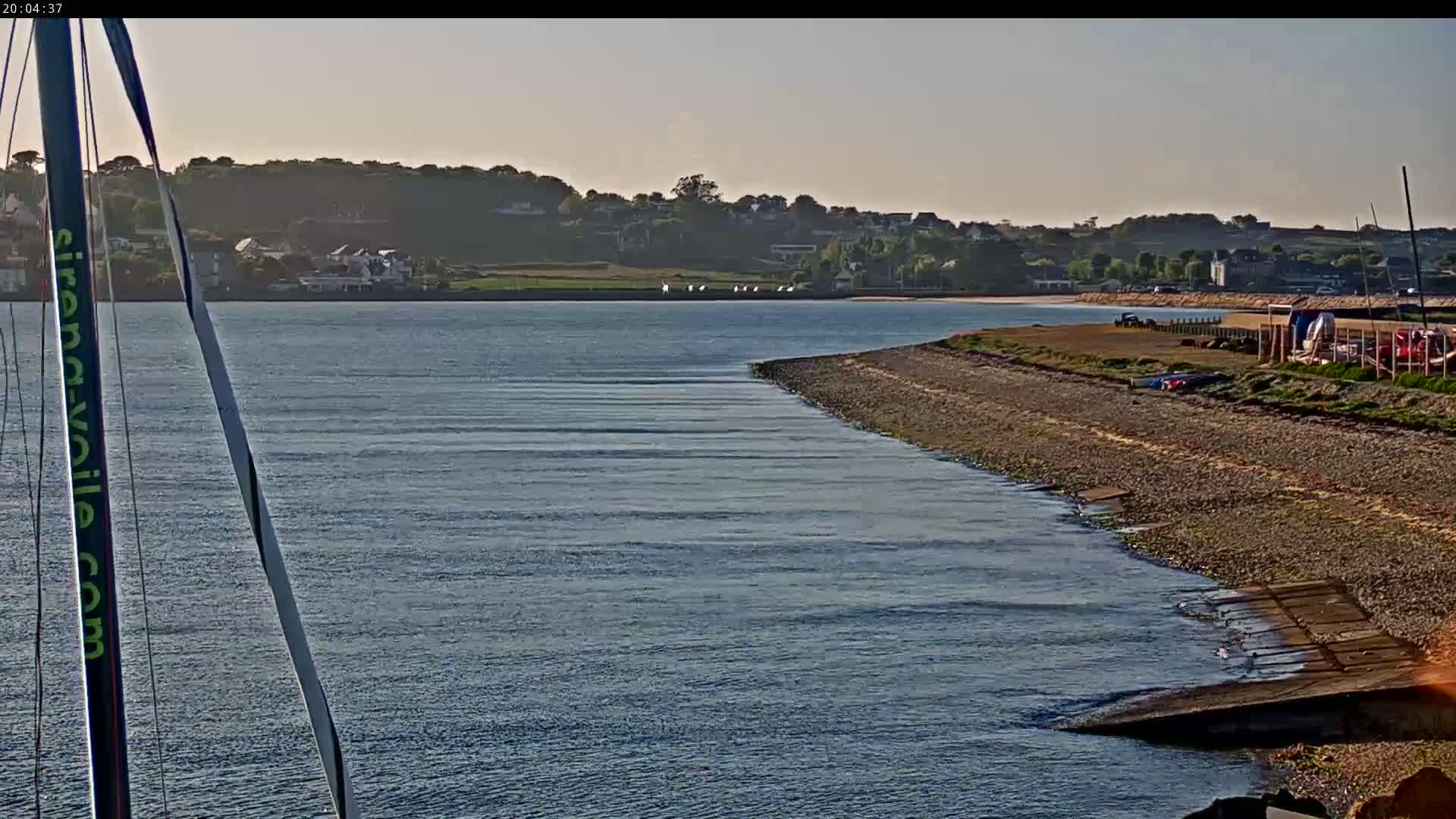 A calm body of water laps a gently sloping, rocky shoreline that leads to a distant, hilly landscape under a clear sky.