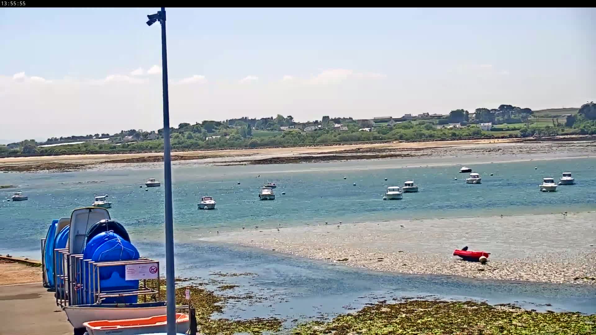 A sunny day shows several small boats moored in calm water near a sandy shore and a distant village.