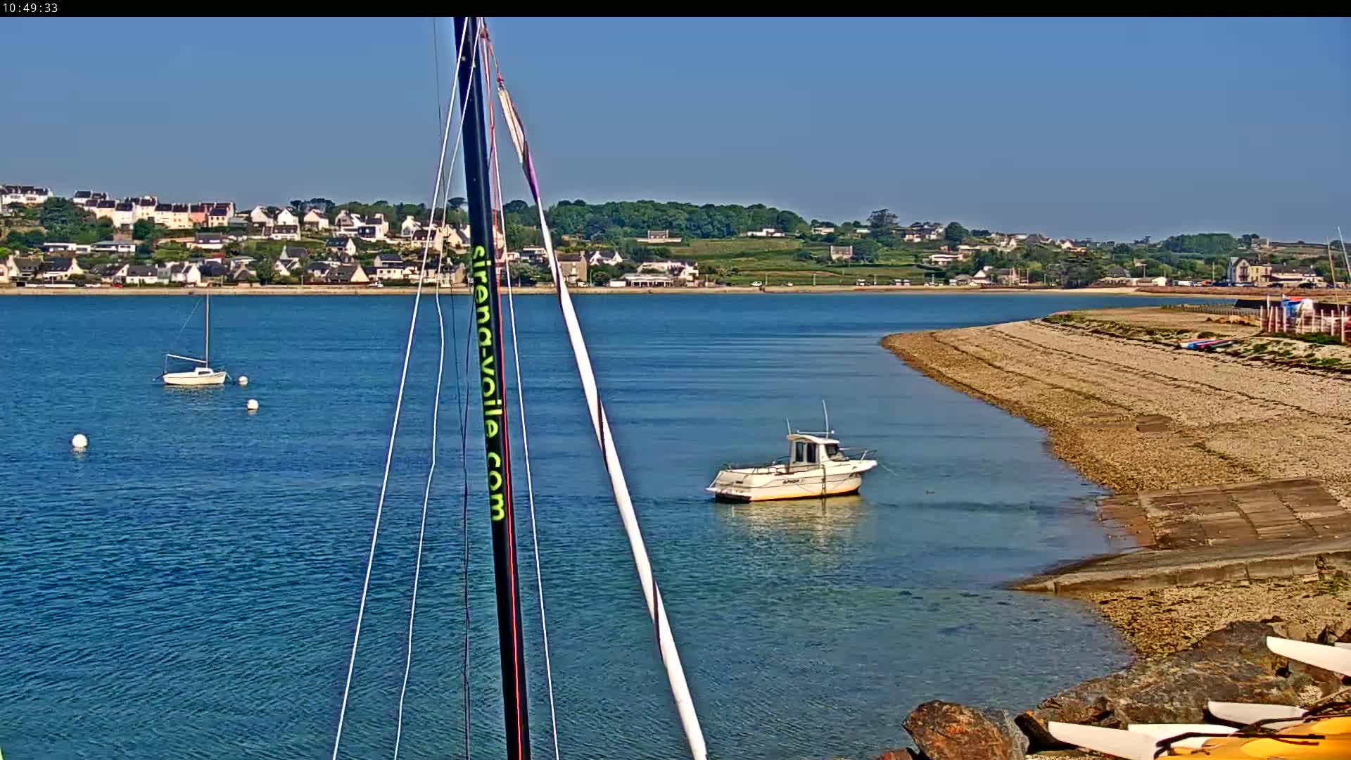 A calm body of water shows a small sailboat and motorboat near a rocky shore, with a village visible in the background on a sunny day.