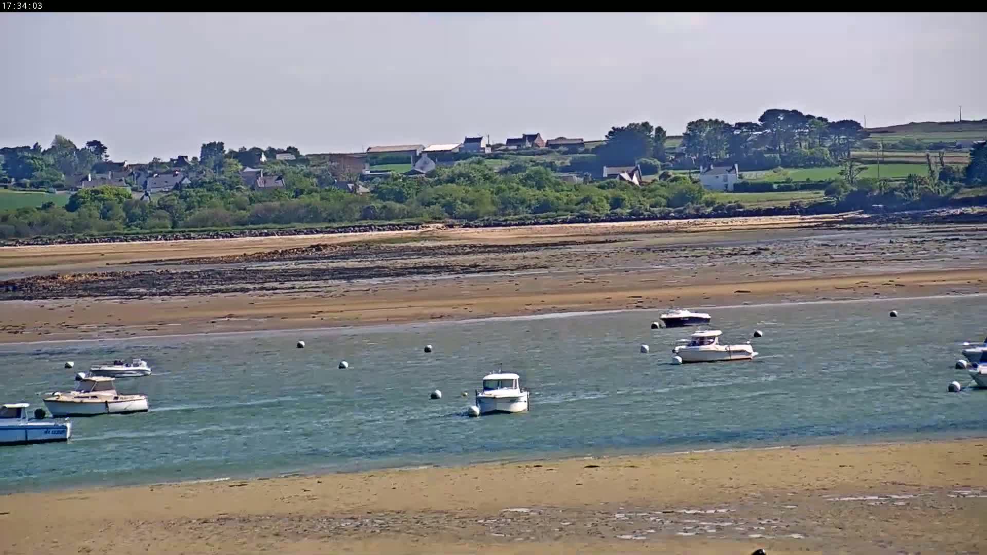 Several small motorboats are moored in calm, shallow water near a sandy shoreline, with a village and green hills visible in the distance under a clear sky.