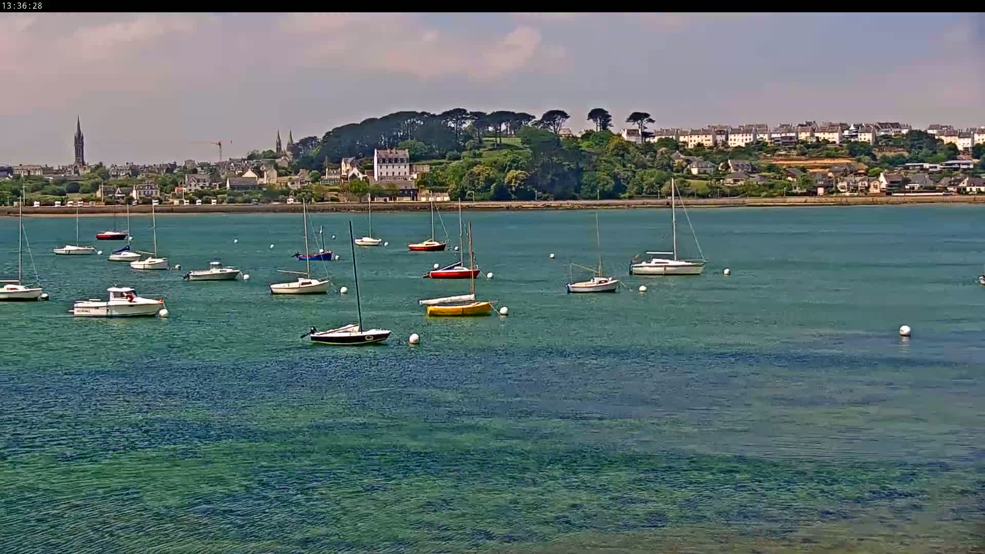 Numerous sailboats and motorboats are moored in calm, clear teal water in front of a coastal town under a mostly sunny sky.