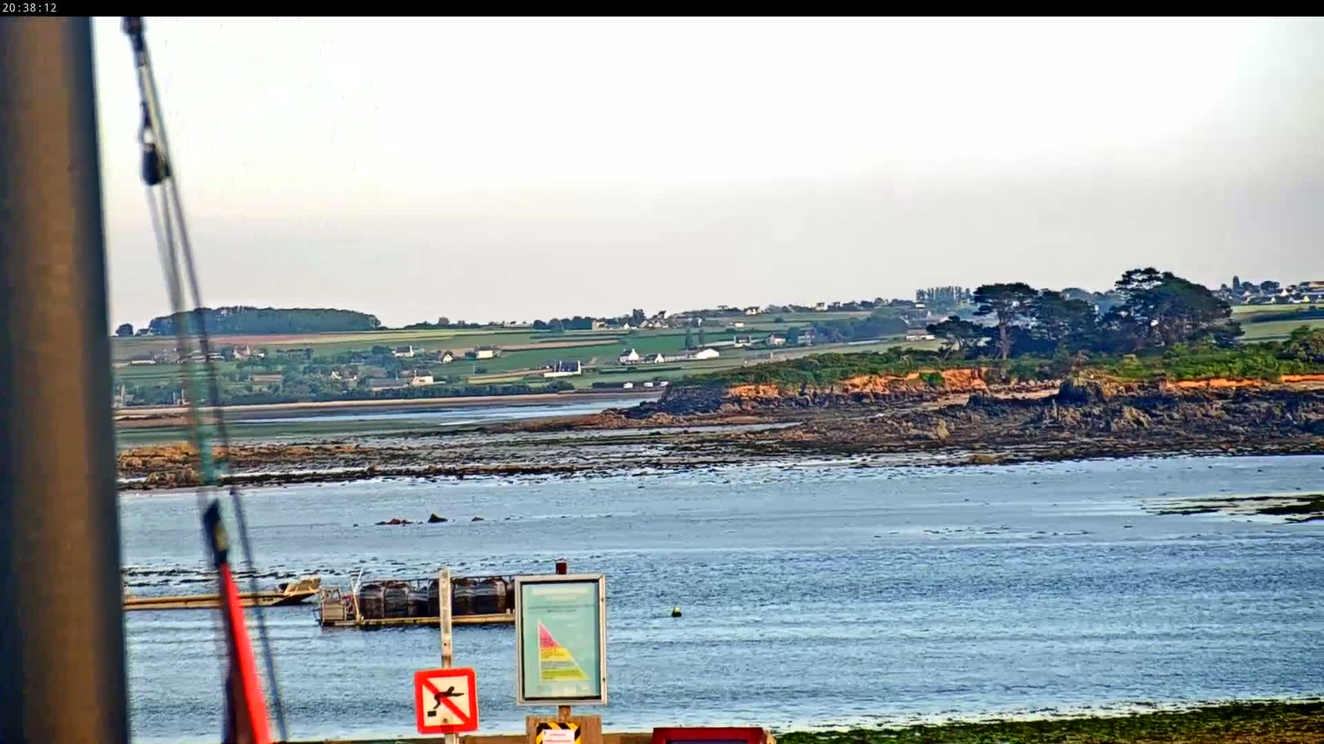A calm body of water stretches out to a rocky shoreline and a verdant landscape dotted with houses under a bright, mostly clear sky.