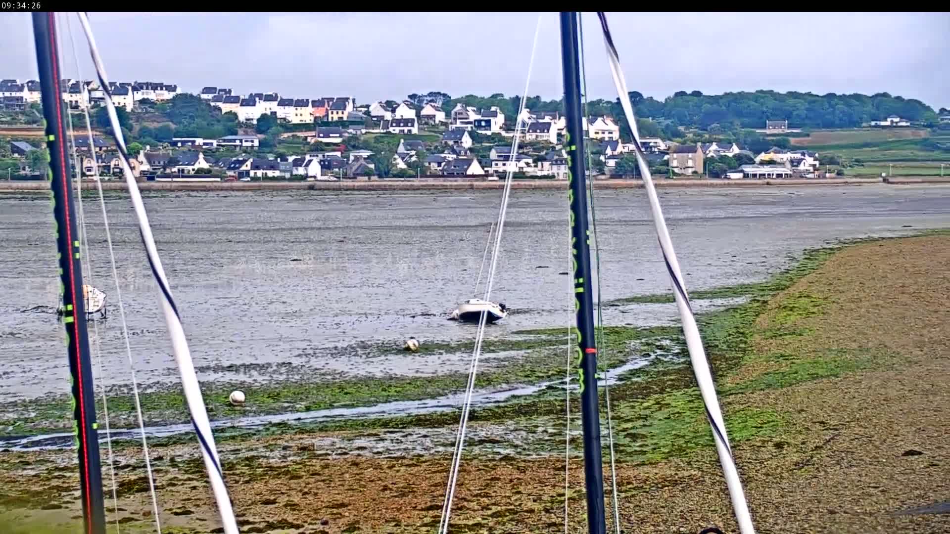 A sailboat is grounded on a mudflat at low tide near a village of white houses, under an overcast sky.