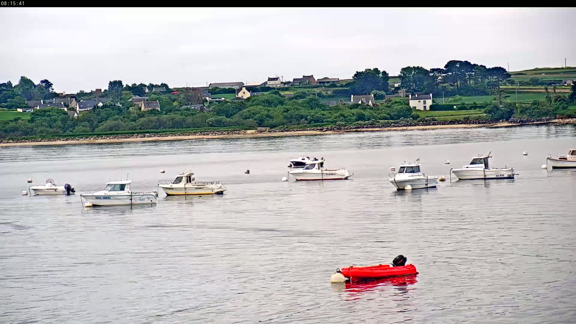 Several small motorboats are moored in calm water in front of a coastal village under an overcast sky.
