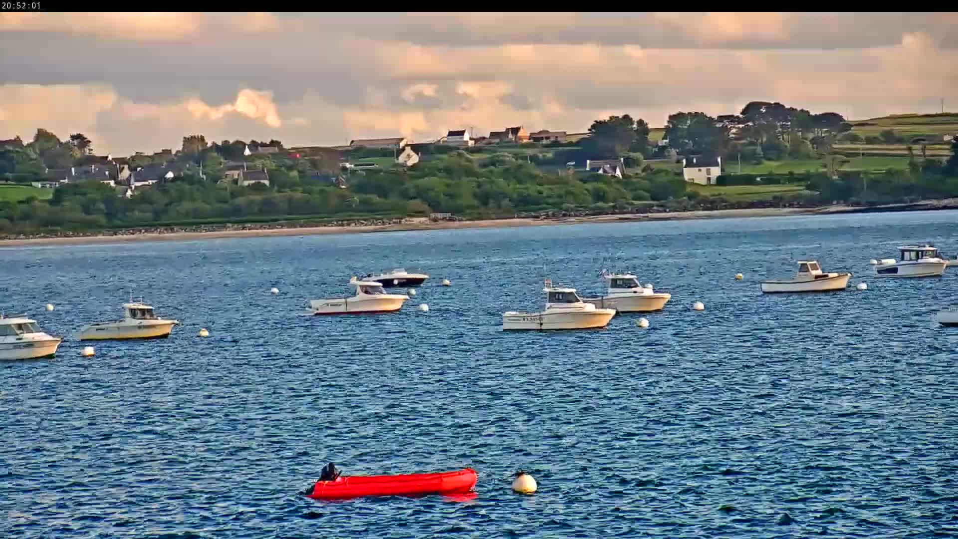 Several small motorboats are moored in calm blue water near a shoreline with houses and green hills under a partly cloudy sky.