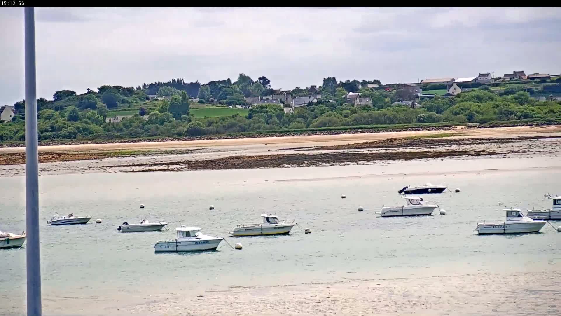Several small motorboats are moored in calm, shallow water near a sandy shore, with a village and green hills visible in the background under a partly cloudy sky.