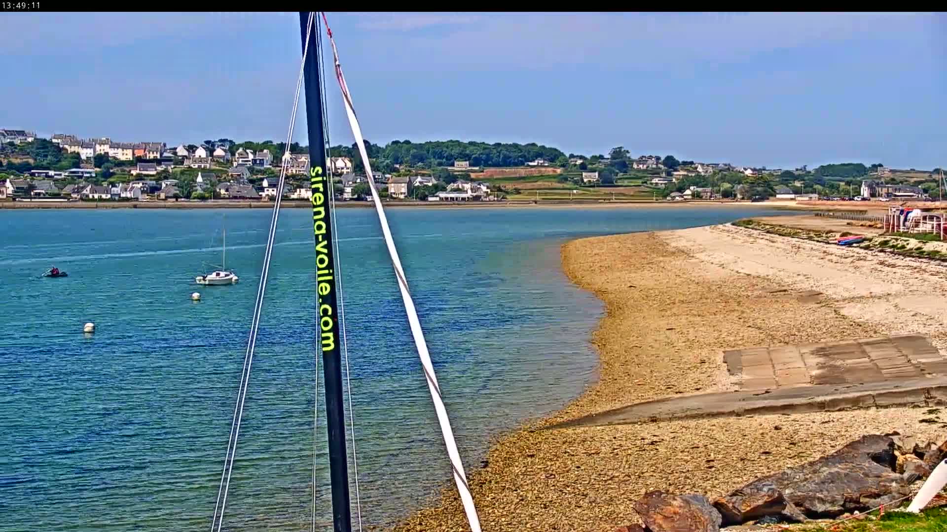 A sailboat mast is in the foreground, overlooking a calm bay with a sandy beach and a small town in the distance under a clear blue sky.